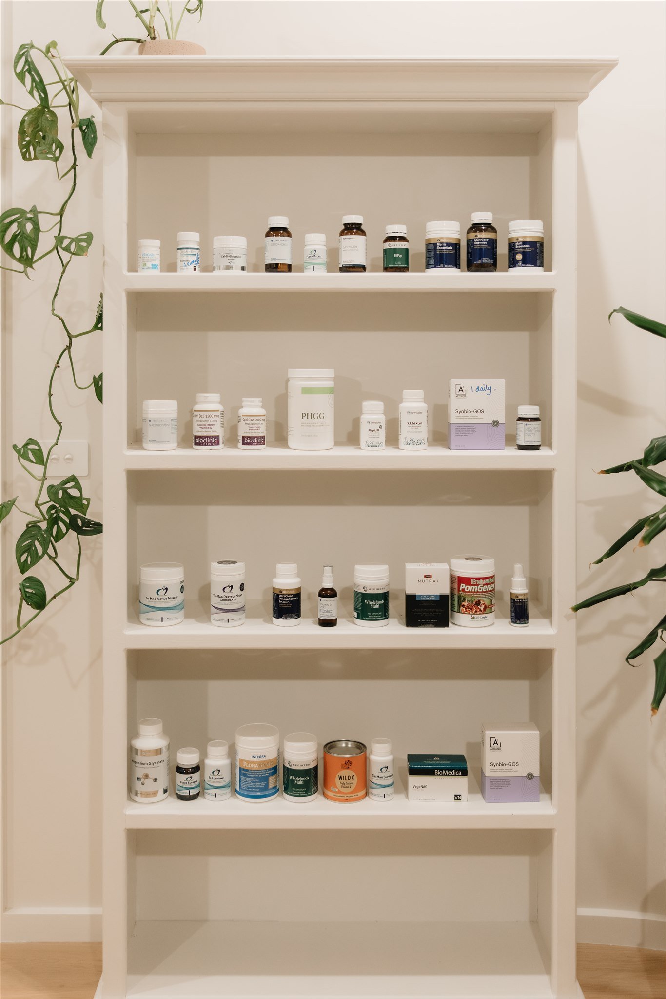 A white bookshelf with five shelves holding various supplement bottles and boxes, with plants on either side. The top shelf has small bottles, the second shelf has a mix of bottles and a box, the third shelf has larger bottles and a box, the fourth shelf has various supplement bottles, and the bottom shelf has more bottles and boxes.