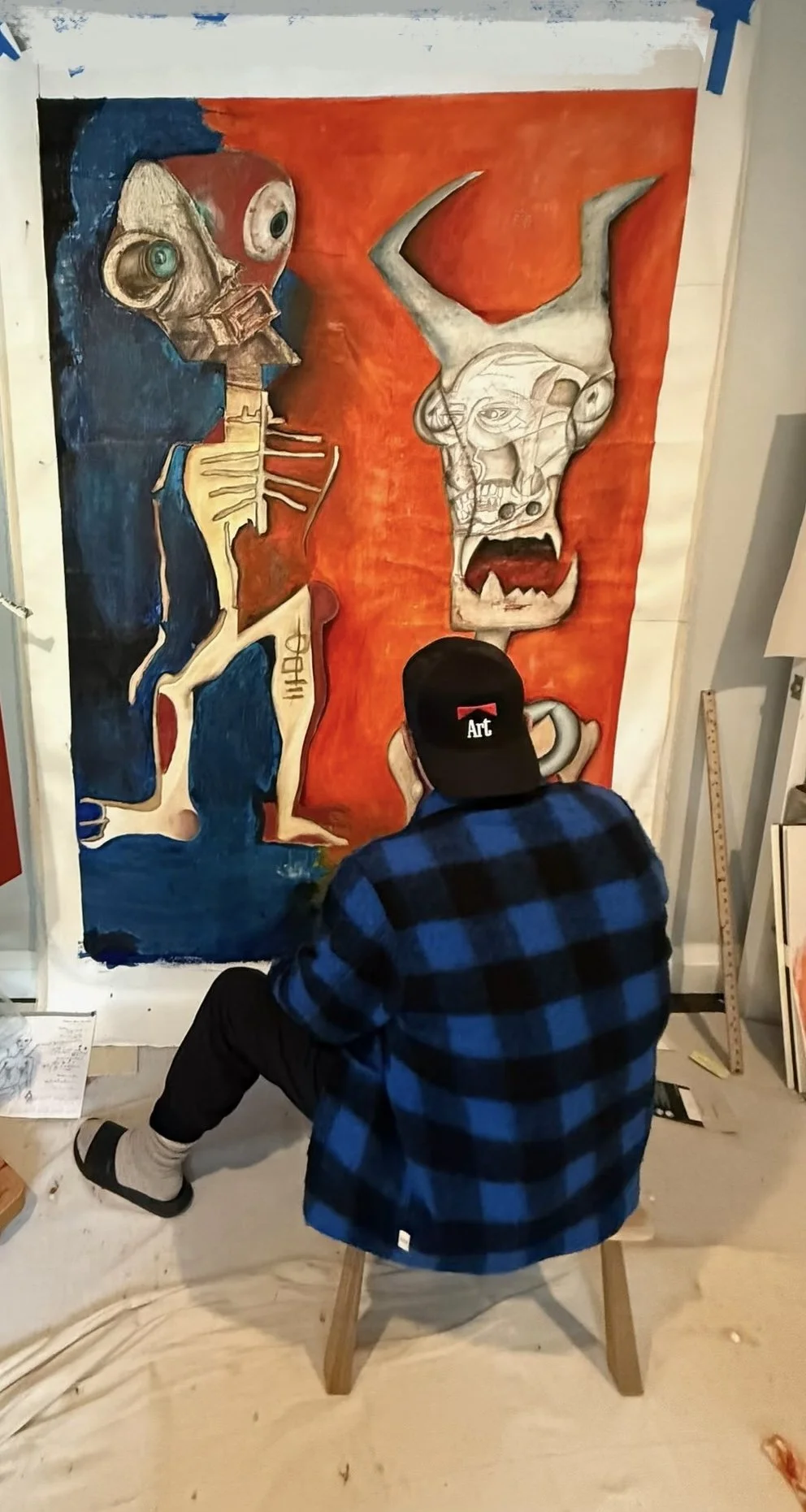 Ron Aniello seated on a stool in his studio, seen from behind, working on a large unstretched canvas of two horned, skeletal figures in red and blue. The artist at work on a Neo-Expressionist painting.