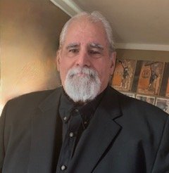 Portrait of a middle-aged man with a gray beard and gray hair, wearing a black bunnet and black suit, standing indoors in front of a wall with decorative framed pictures.