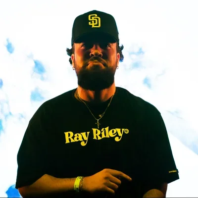 A young man wearing a black baseball cap with yellow logo, black t-shirt with yellow text 'Ray Riley', gold necklace, and yellow wristband, standing against a cloudy sky background.