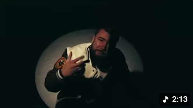 A man taking a selfie in front of a dark background, illuminated by a circular light, wearing a jacket with patches, making a peace sign and smiling.