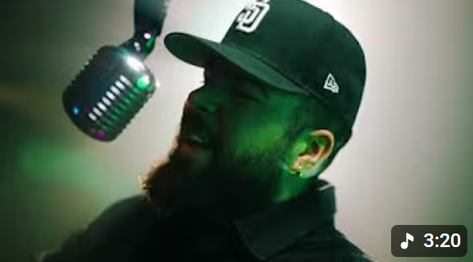 A man with a beard wearing a cap and headphones singing into a microphone in a recording studio with green light.