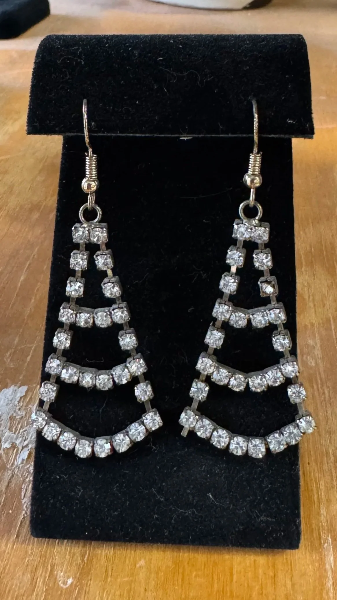 A pair of silver dangling earrings with multiple rows of clear rhinestones displayed on a black velvet stand.