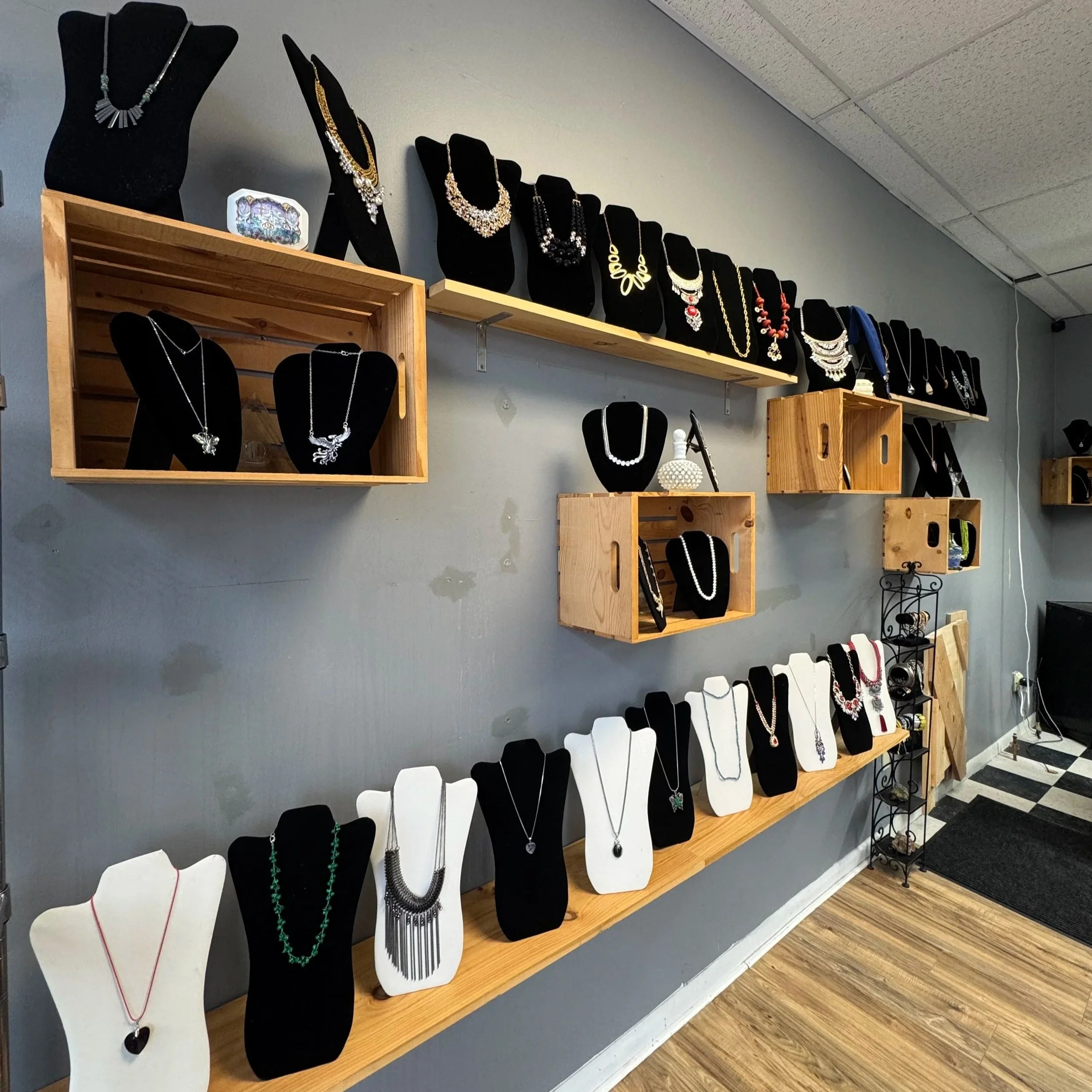Display of various necklaces on black and white busts and in wooden and metal shelves against a gray wall.
