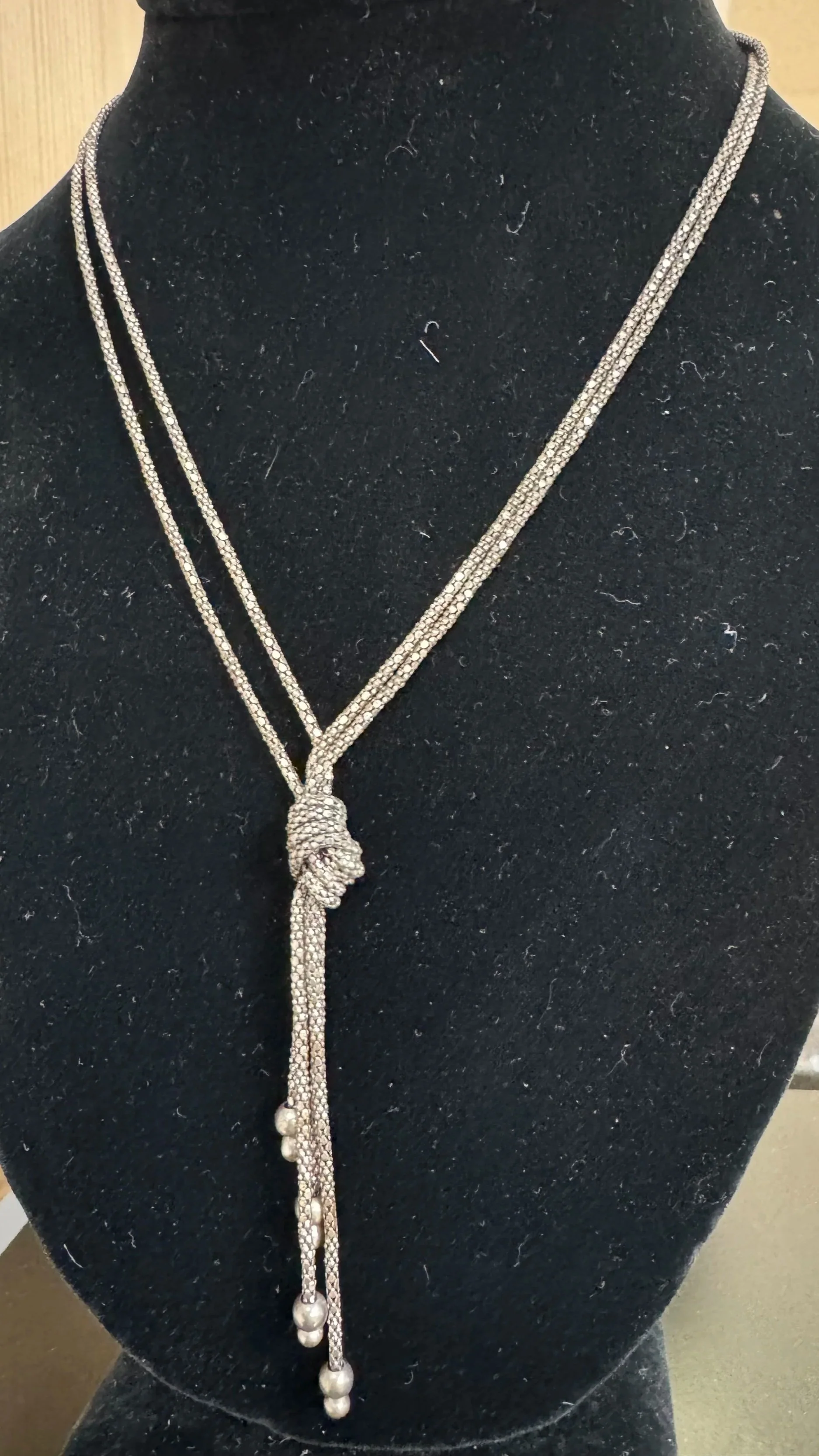 A silver, diamond-encrusted necklace with a knot detail in the center and dangling pearl accents, displayed on a black velvet bust.