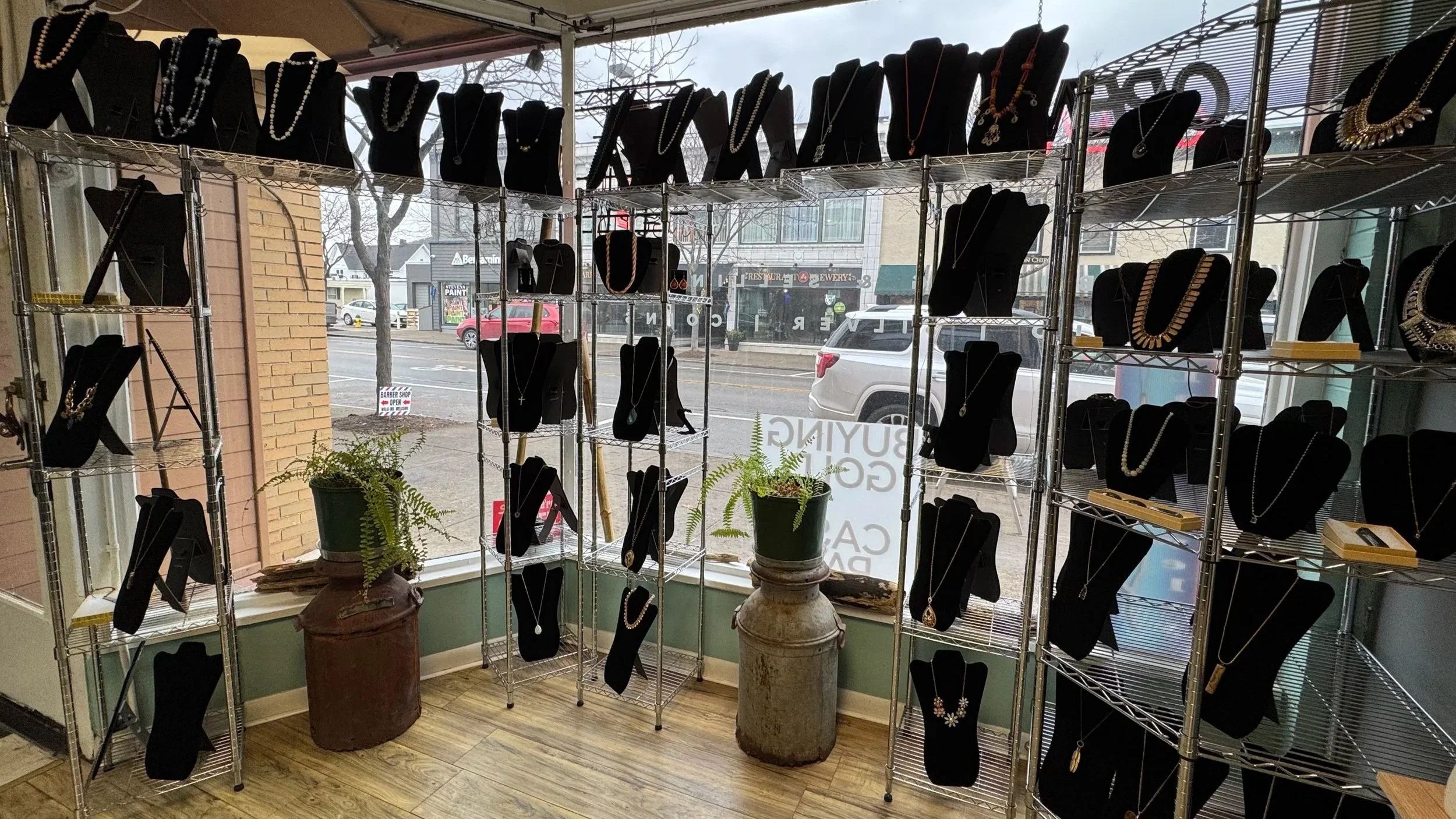Jewelry store display with necklaces on black busts on metal shelves inside storefront, front window view of street with parked cars and buildings outside.