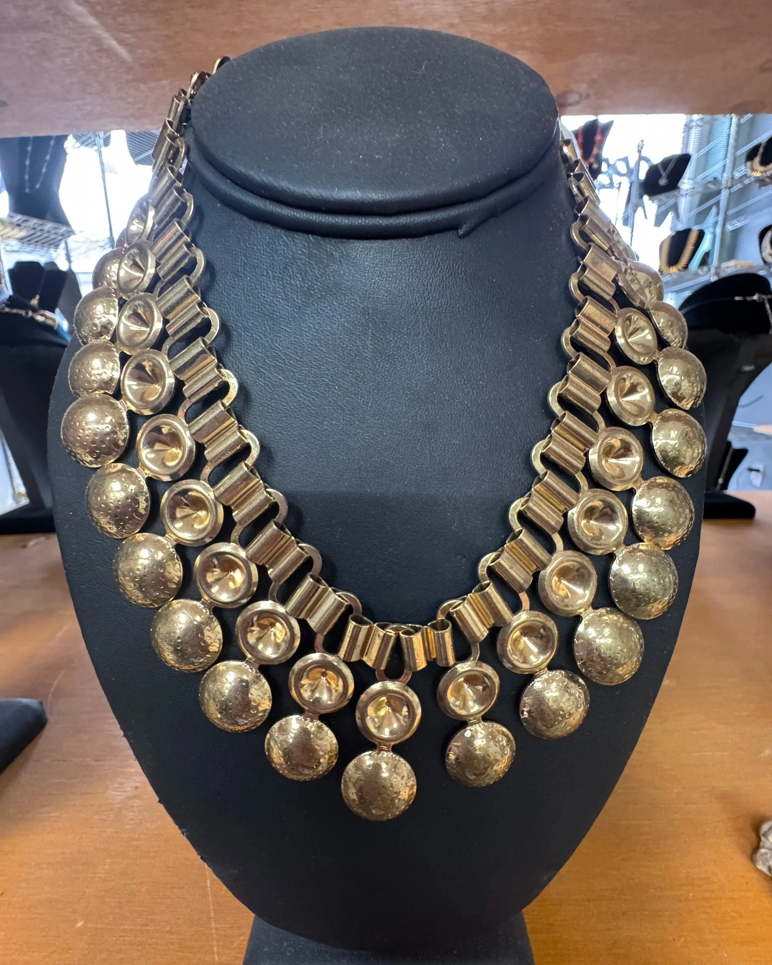 A gold statement necklace with large, textured, circular and geometric links displayed on a black jewelry stand.