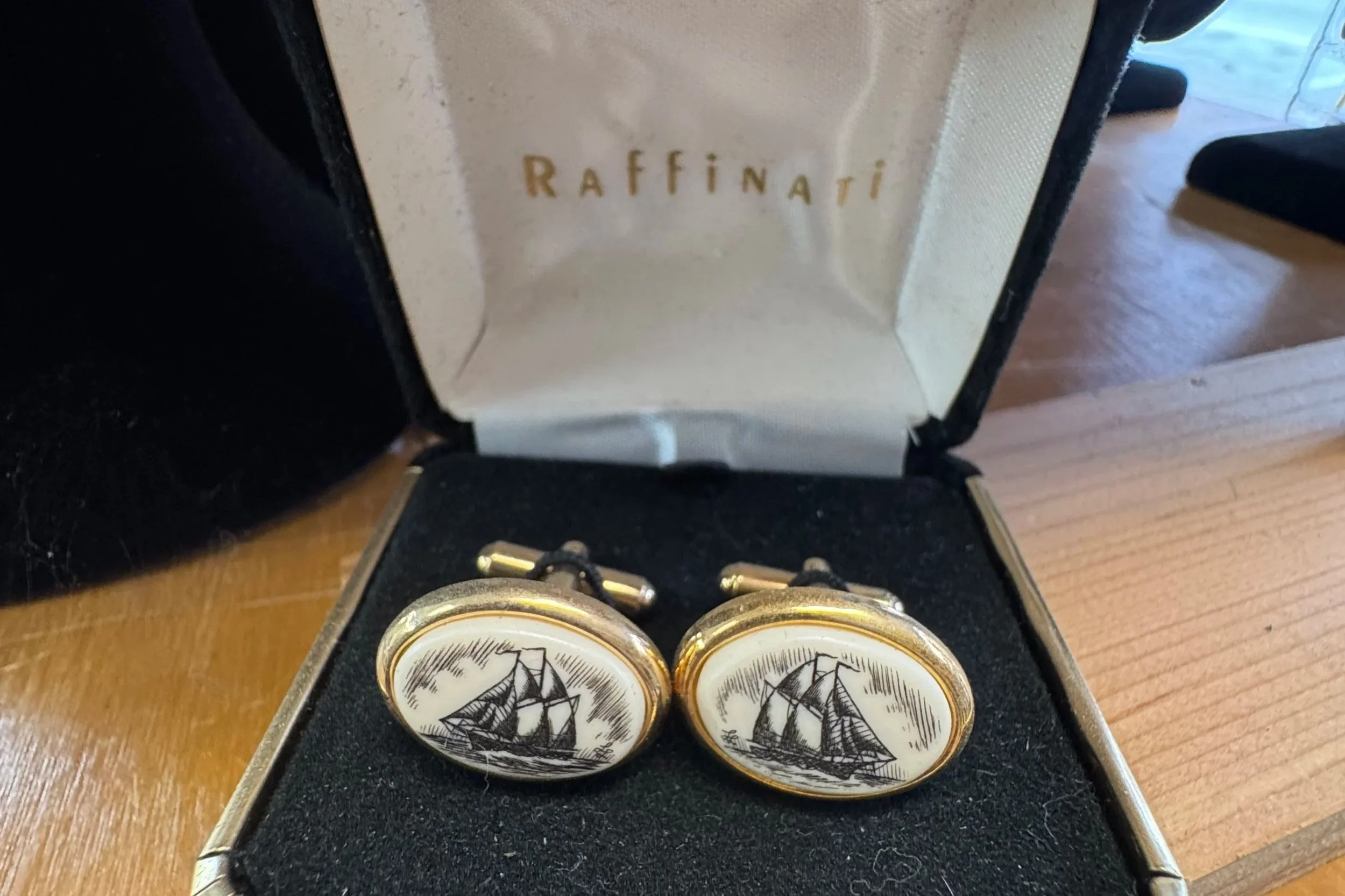 Gold cufflinks with a black and white sailing ship illustration in a black velvet-lined box, with the brand name 'Raffinati' inside the box lid.