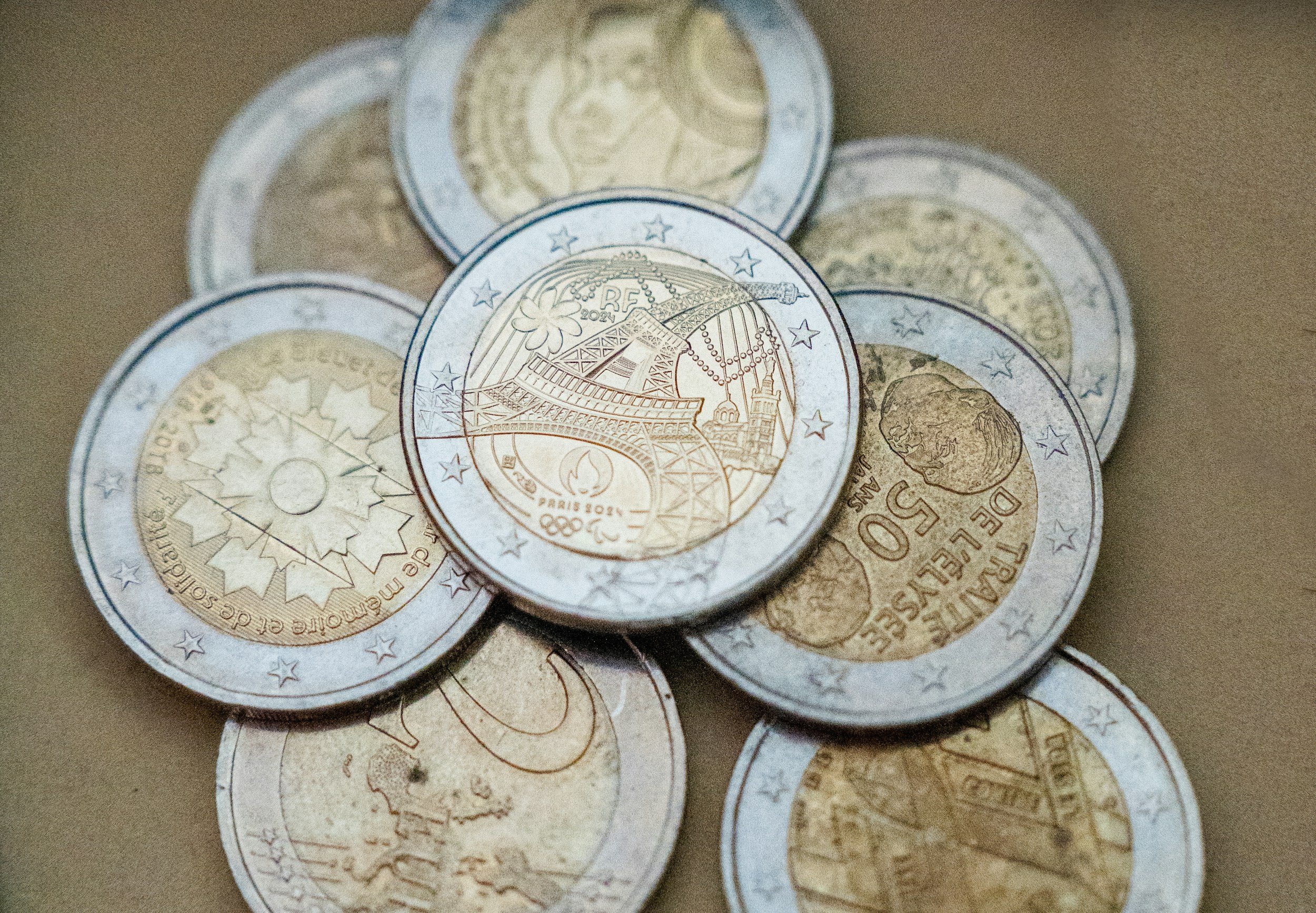 A pile of euro coins, including a 2 euro coin with a depiction of the Eiffel Tower and a flower, a 1 euro coin, and other coins with various designs, on a brown surface.