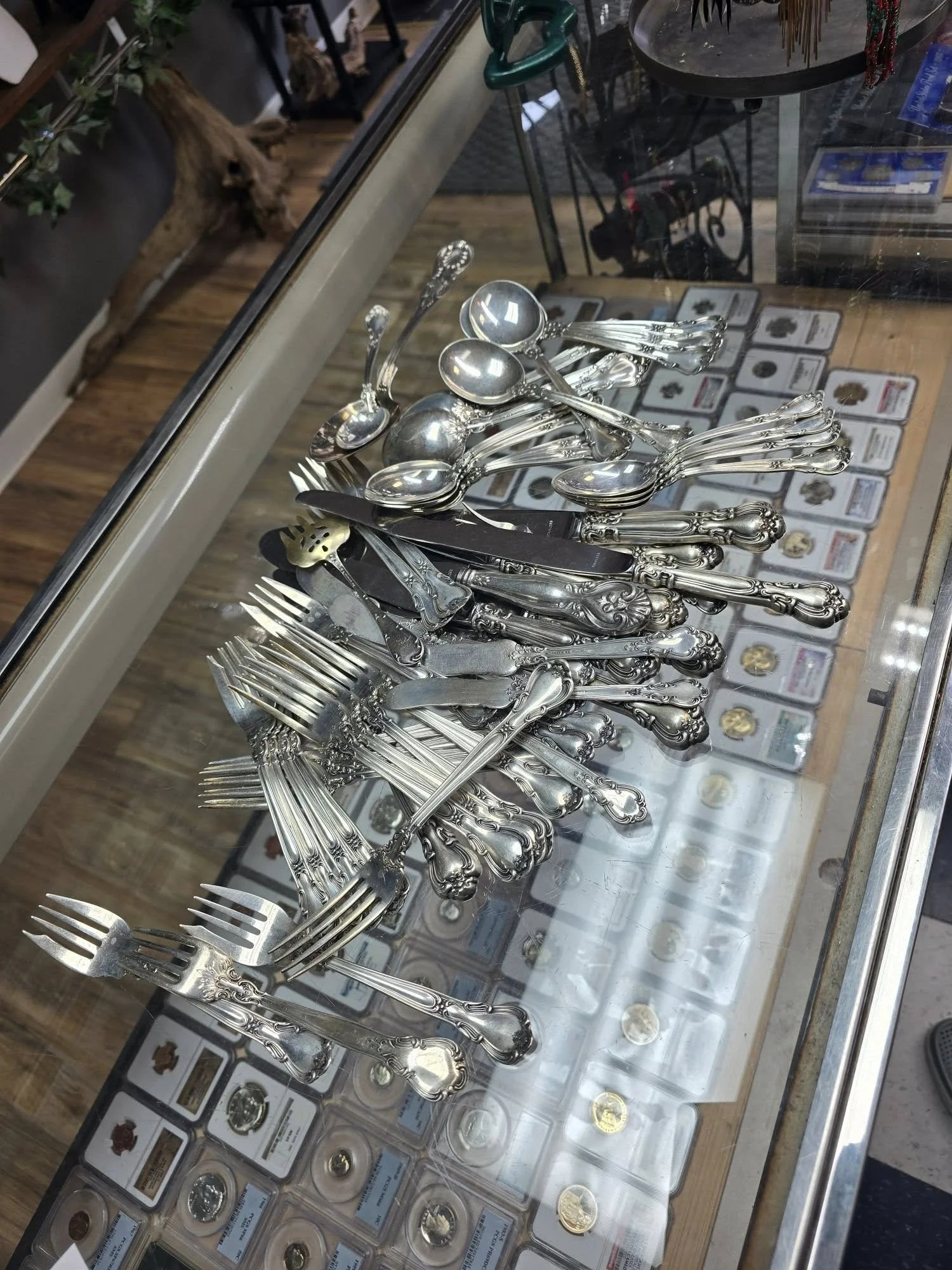 Assorted silverware including forks, knives, and spoons displayed on a glass table with coins and collectibles beneath.