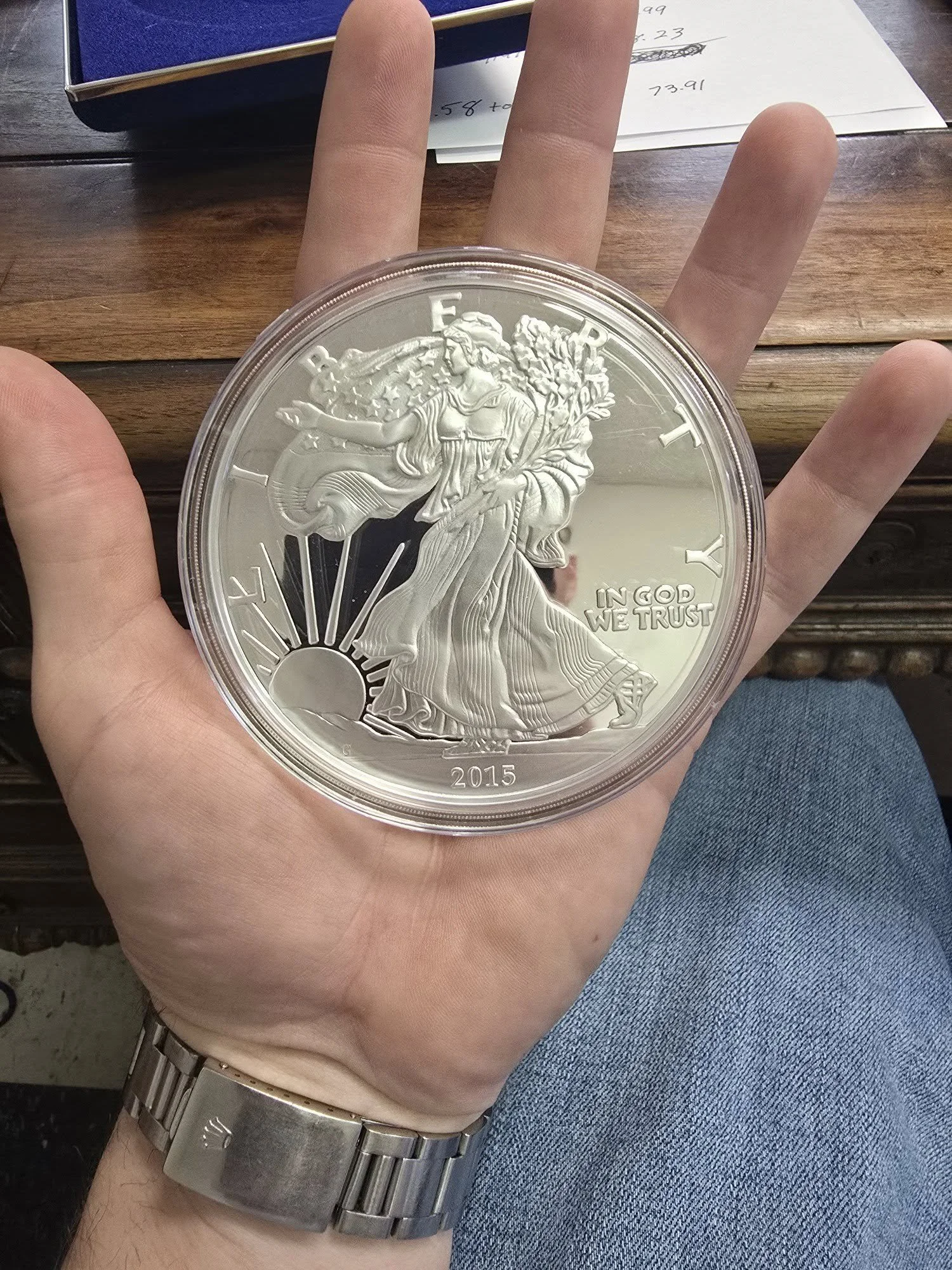 A person's hand holding a large silver coin with an engraved image of Lady Liberty, rays of sunlight, and the year 2015, on a wooden table.