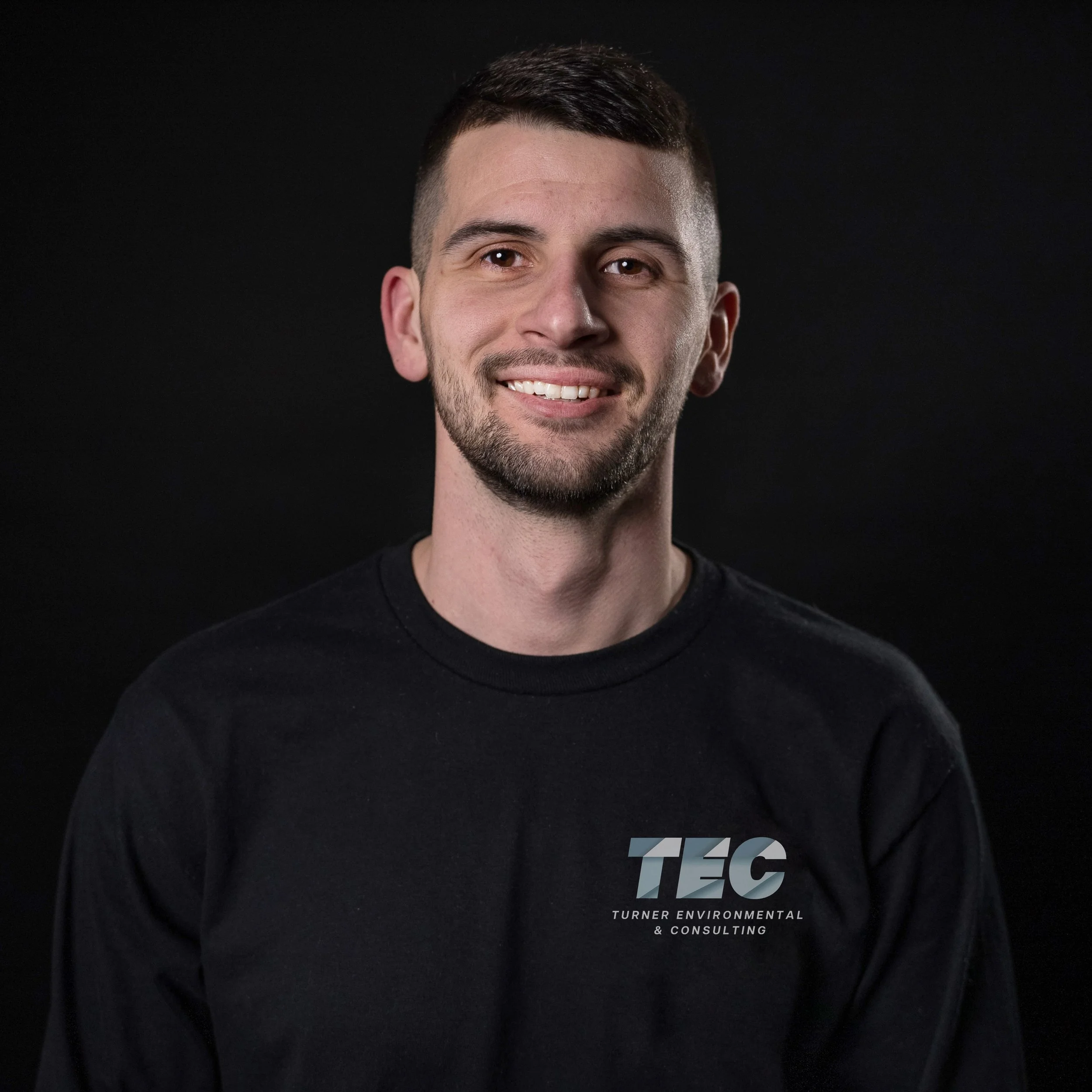 A young man with short dark hair and a beard, smiling, wearing a black T-shirt with the logo 'T.E.C' and the text 'Turner Environmental & Consulting' on the front, against a black background.