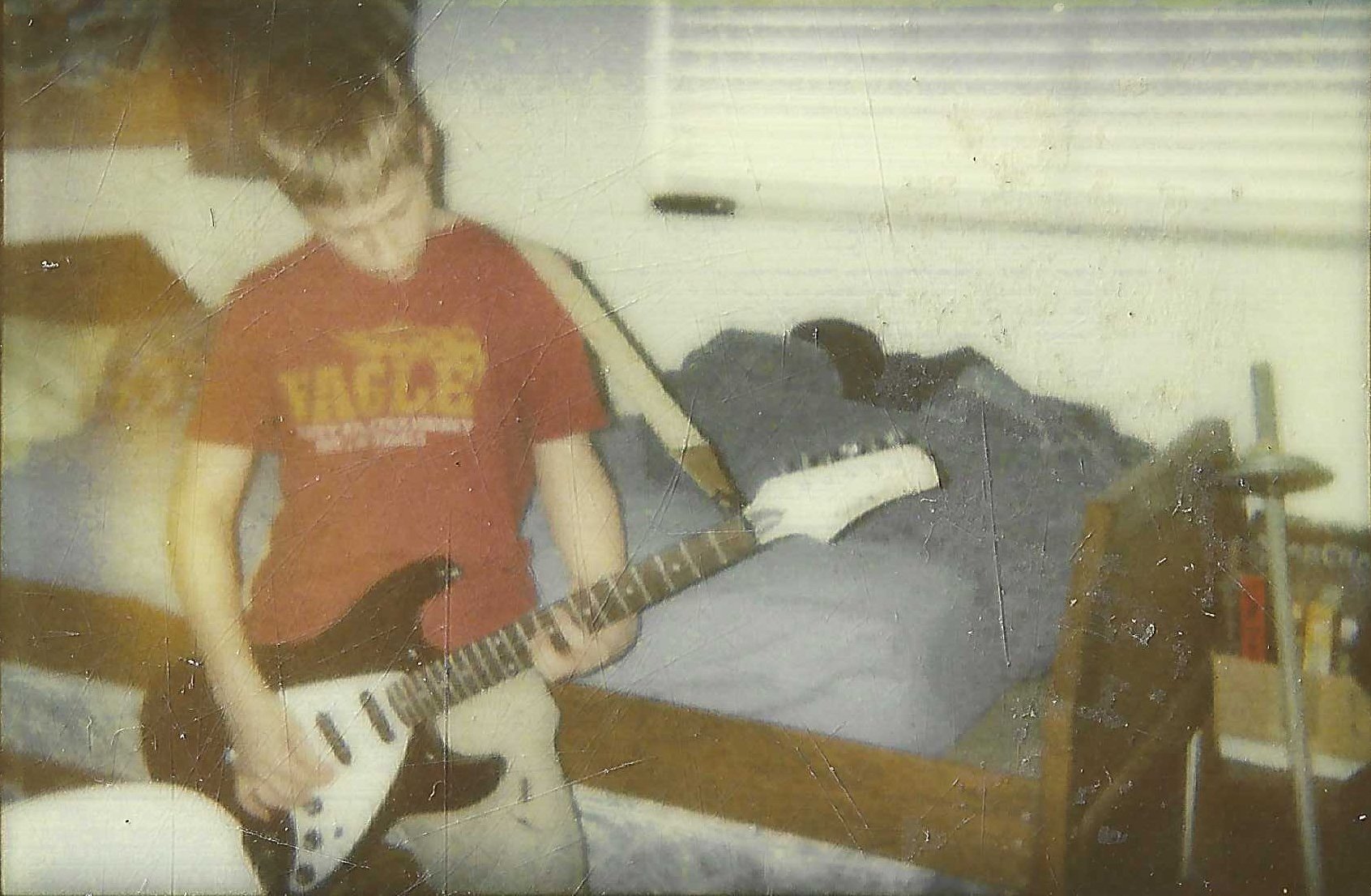 A young boy with a guitar stands near a bed. The boy appears to be playing the guitar in a dimly lit room.