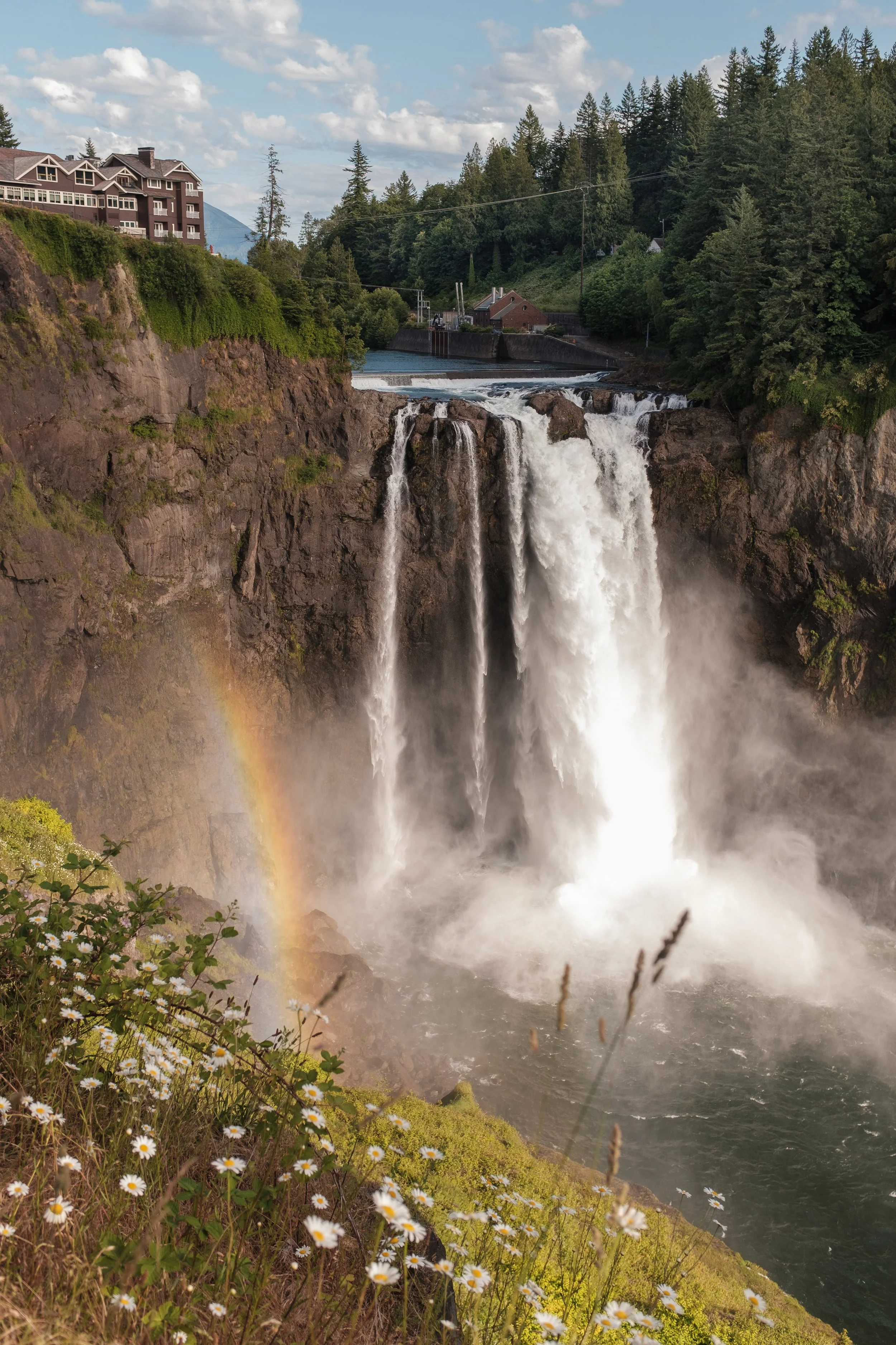 Waterfalls with mist and rainbow, greenery in foreground, trees and buildings in background.
