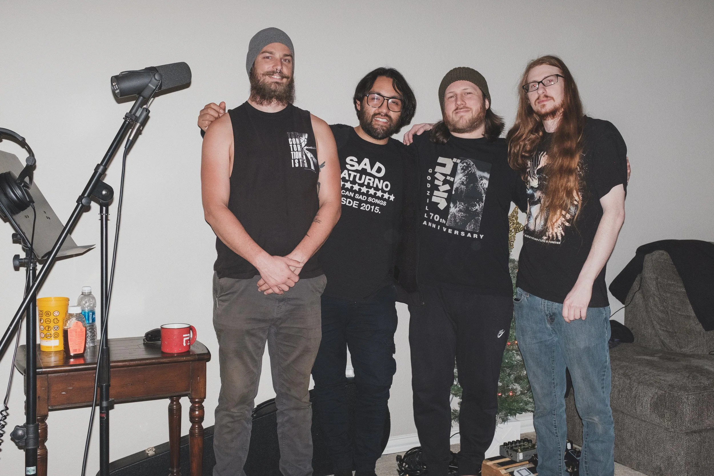 Four men with long hair and beards posing together indoors, with musical equipment and a small decorated Christmas tree in the background.