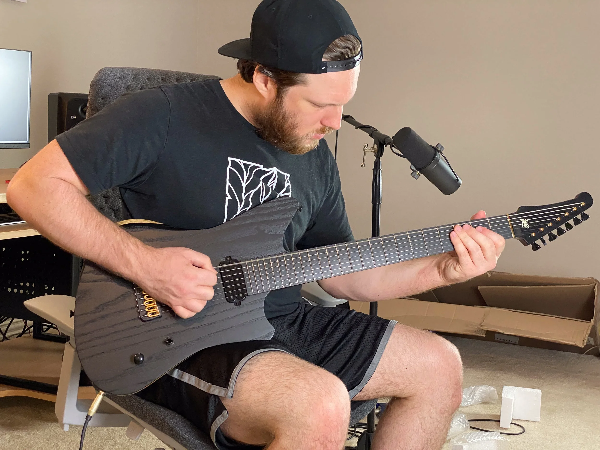 A man wearing a black cap, black t-shirt, and black shorts, sitting on a chair, playing a black electric guitar. He is focused on playing the guitar in what appears to be a room with recording equipment and an open cardboard box on the floor.