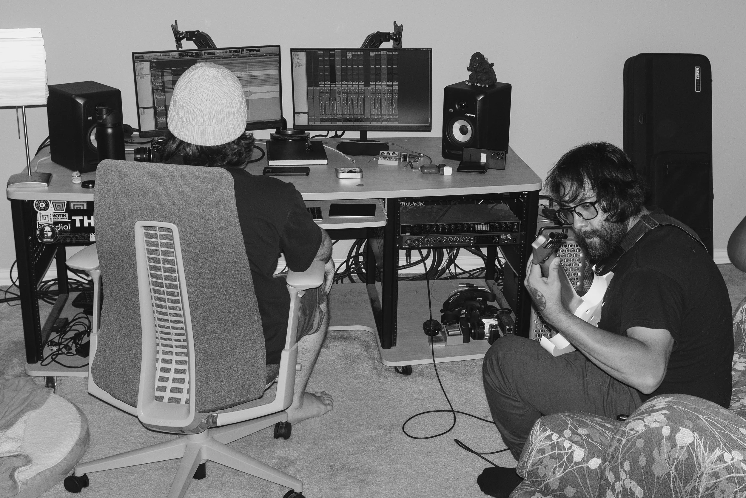 Two young men in a home studio, one seated at a desk with dual monitors and audio equipment, the other sitting in a chair playing an electric guitar.