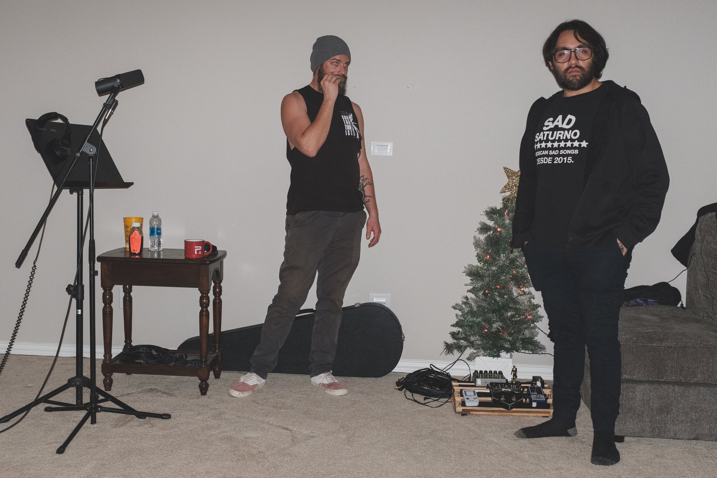 Two men stand in a music studio next to a small decorated Christmas tree, with music equipment and a guitar case nearby.