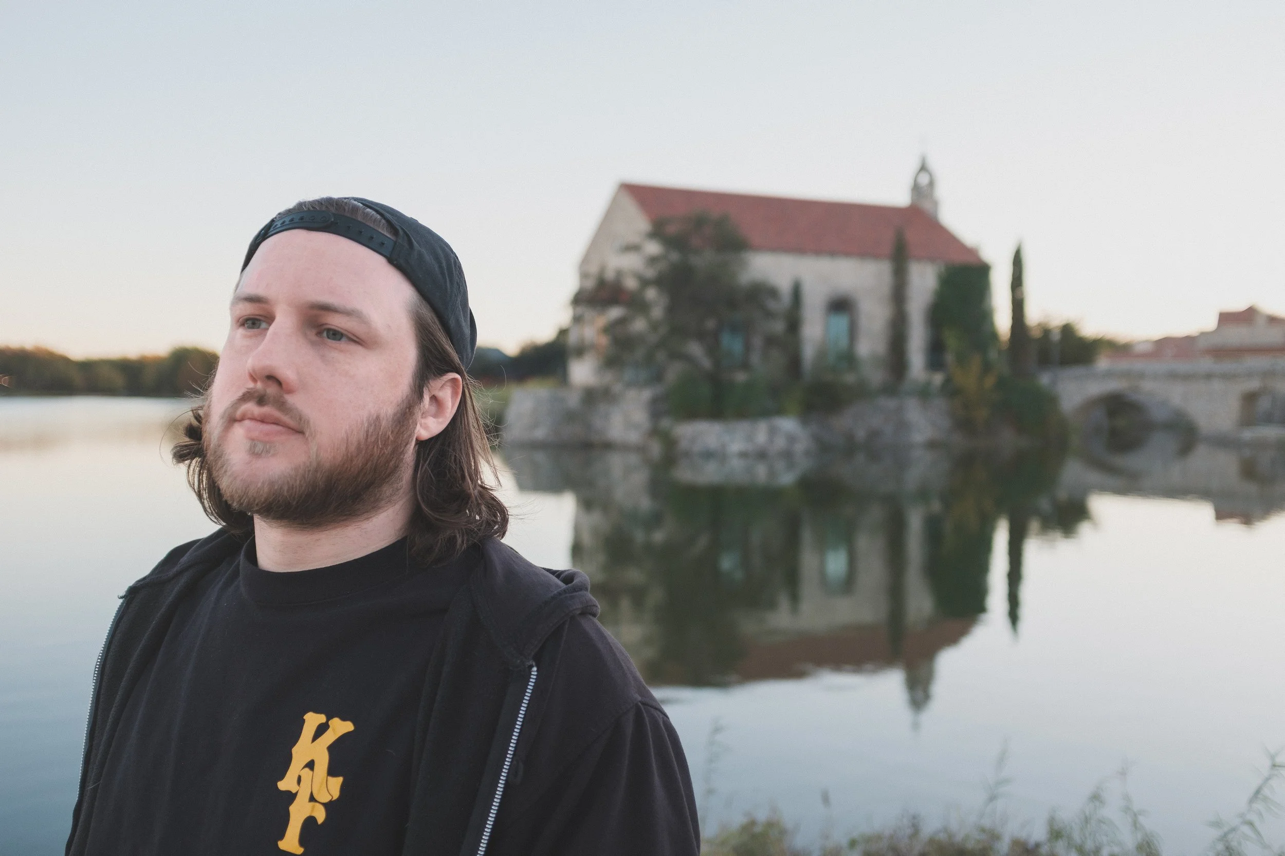 A man with long brown hair, beard, wearing a black baseball cap backward and a black hoodie, standing near a body of water with a historic stone building with red roof and trees reflecting in the water behind him.