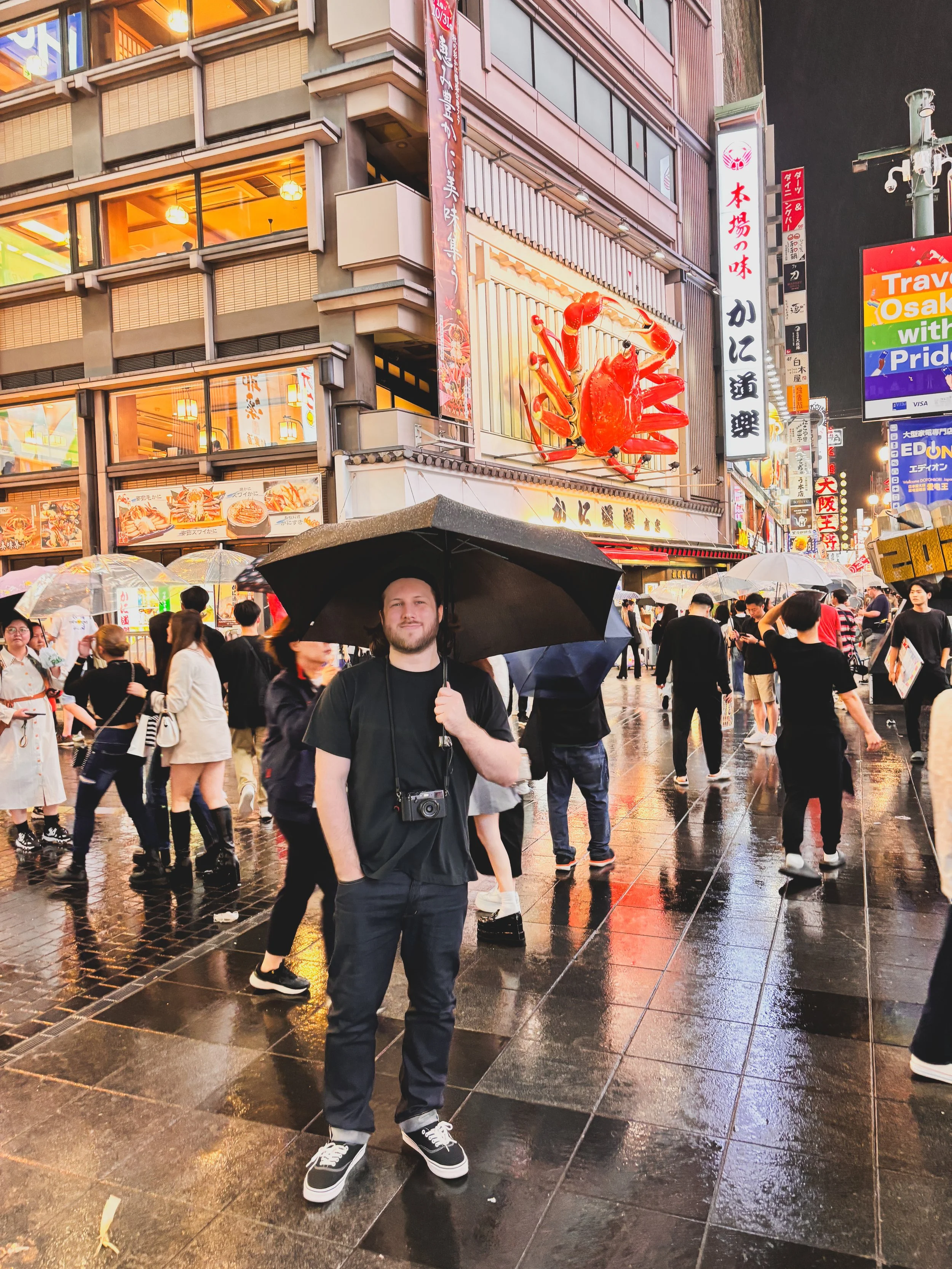 A man holding a black umbrella stands on a wet street in a busy city with neon signs and a large red crab sculpture on a building. The crowd is diverse, and the background features Japanese characters and illuminated storefronts, indicating a lively area possibly in Japan.
