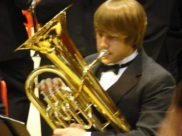 Young man in suit and bow tie playing a brass instrument, likely a euphonium, during a formal event or concert.