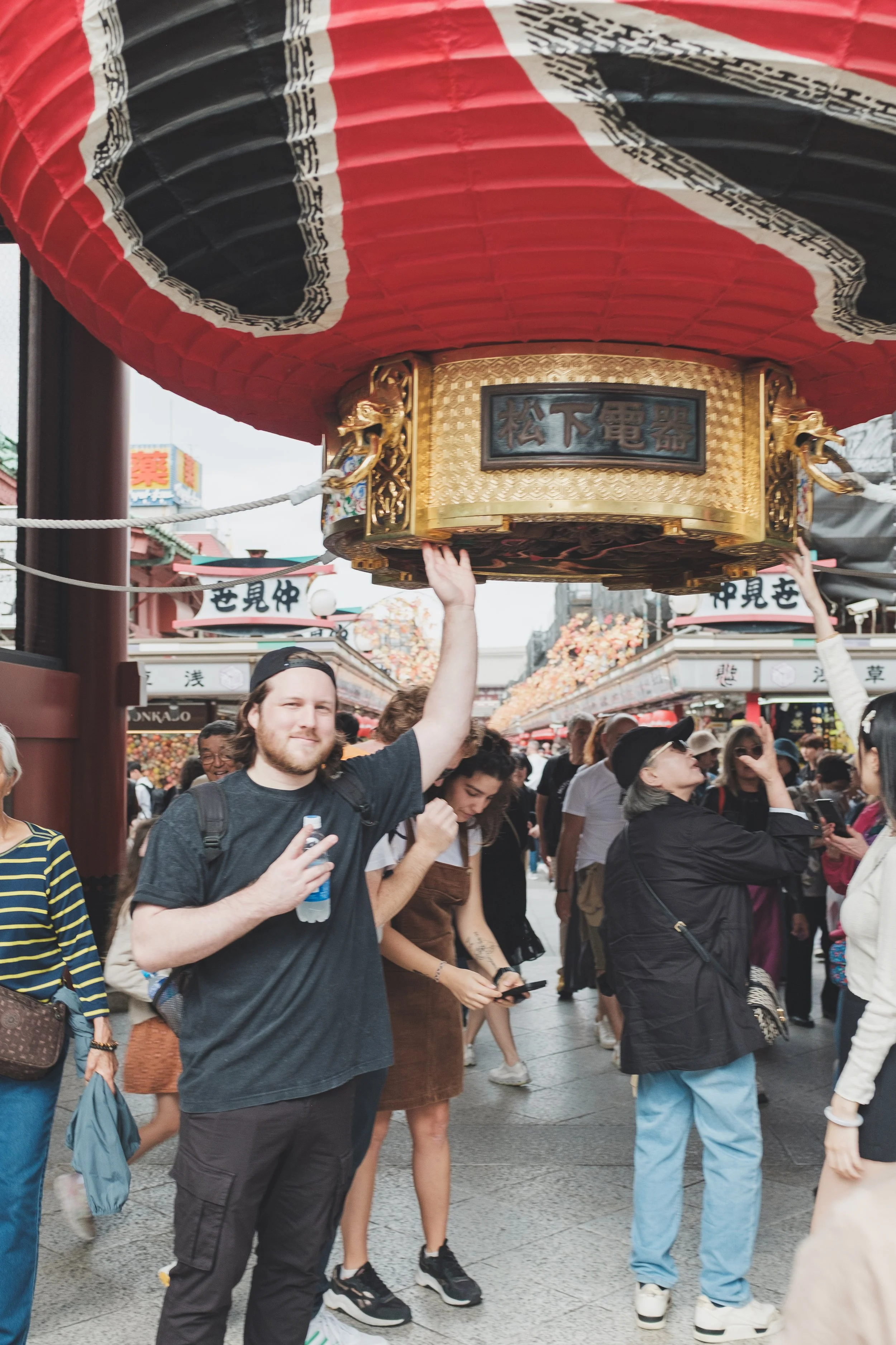 People walking through a busy street at a Japanese festival with a large, decorative red and black paper lantern overhead, featuring Japanese text and gold accents.