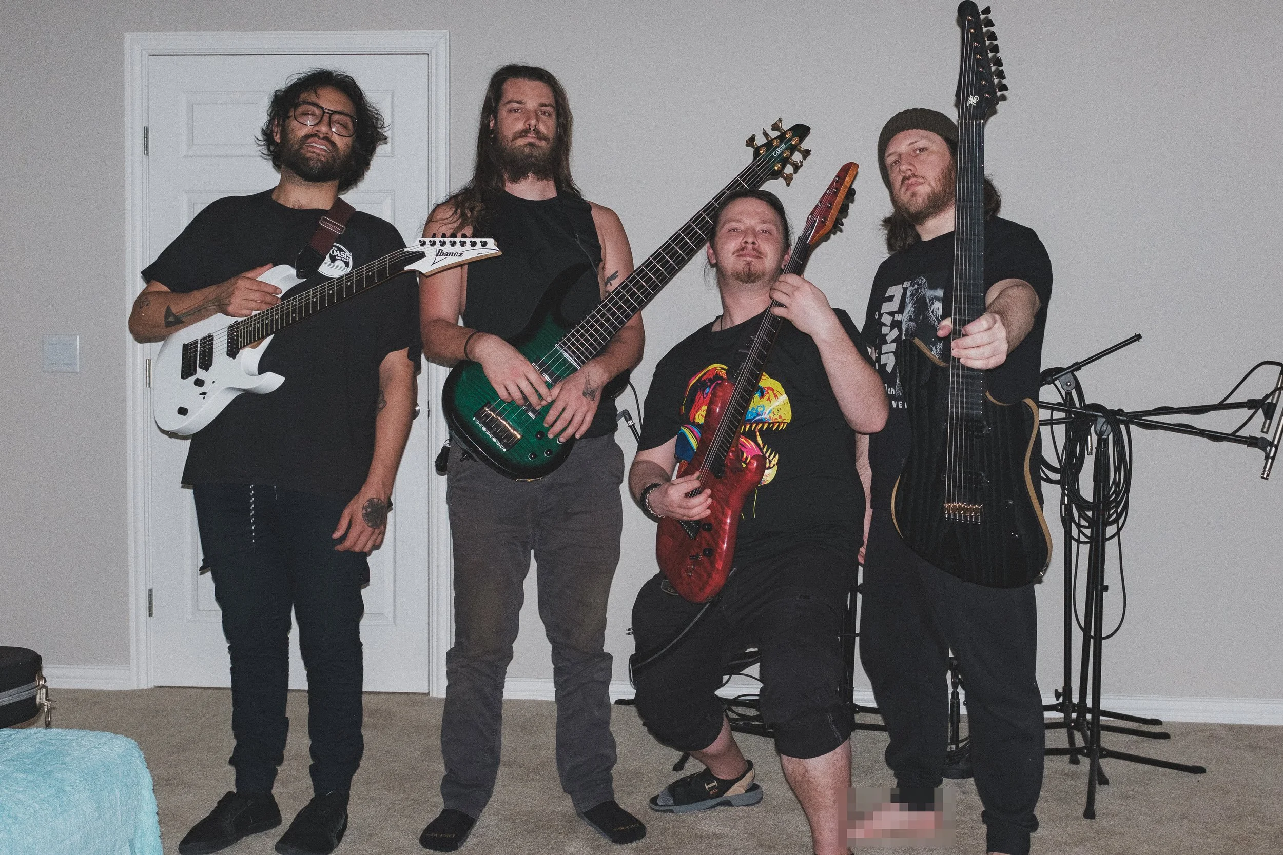 Four men holding electric guitars in a room, with one seated in the middle, smiling and posing for the camera.