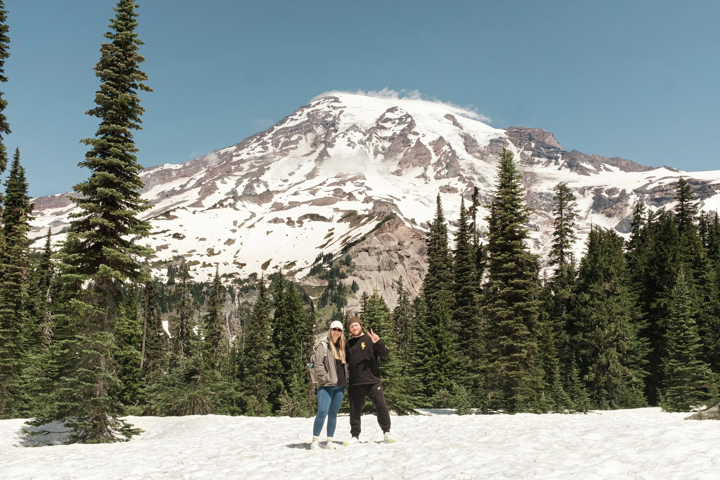 A couple stands on snow with a snow-capped mountain and forest of pine trees in the background.