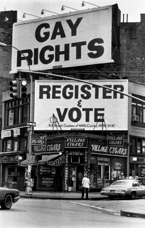 Black and white photo of a busy urban street with two large political signs, one reading "GAY RIGHTS" and the other encouraging people to "REGISTER & VOTE," with several storefronts, signs for "Village Cigars," and a few pedestrians and cars visible.