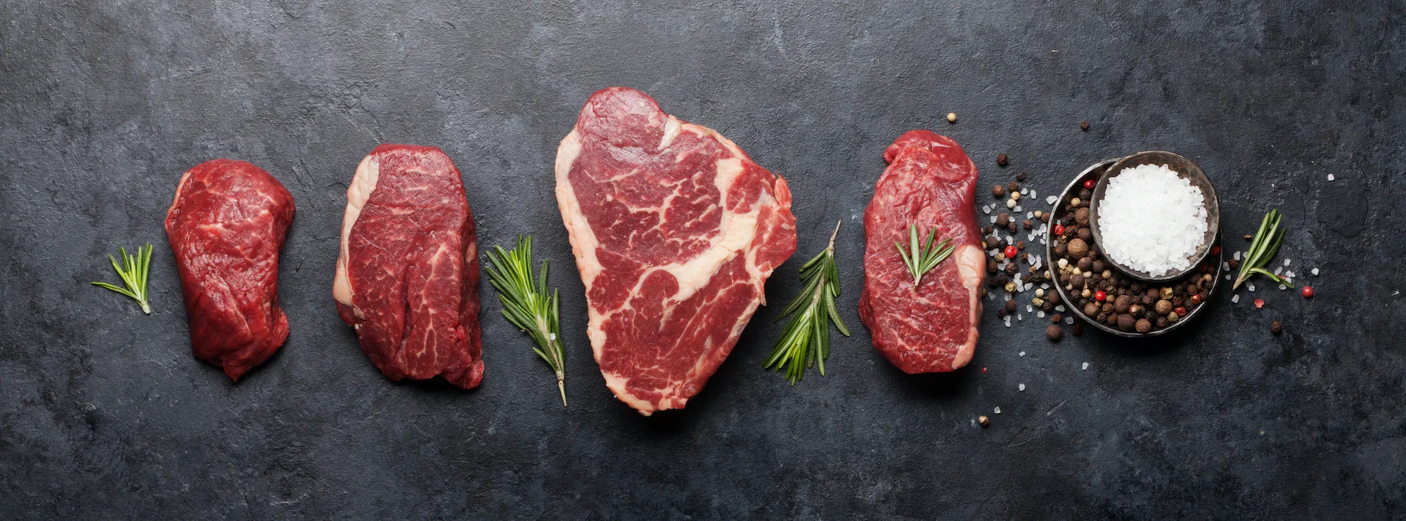 Five pieces of raw red meat with marbling, garnished with sprigs of rosemary, arranged on a dark slate surface. Next to the meat are scattered black, white, and pink peppercorns and a small bowl of coarse sea salt.