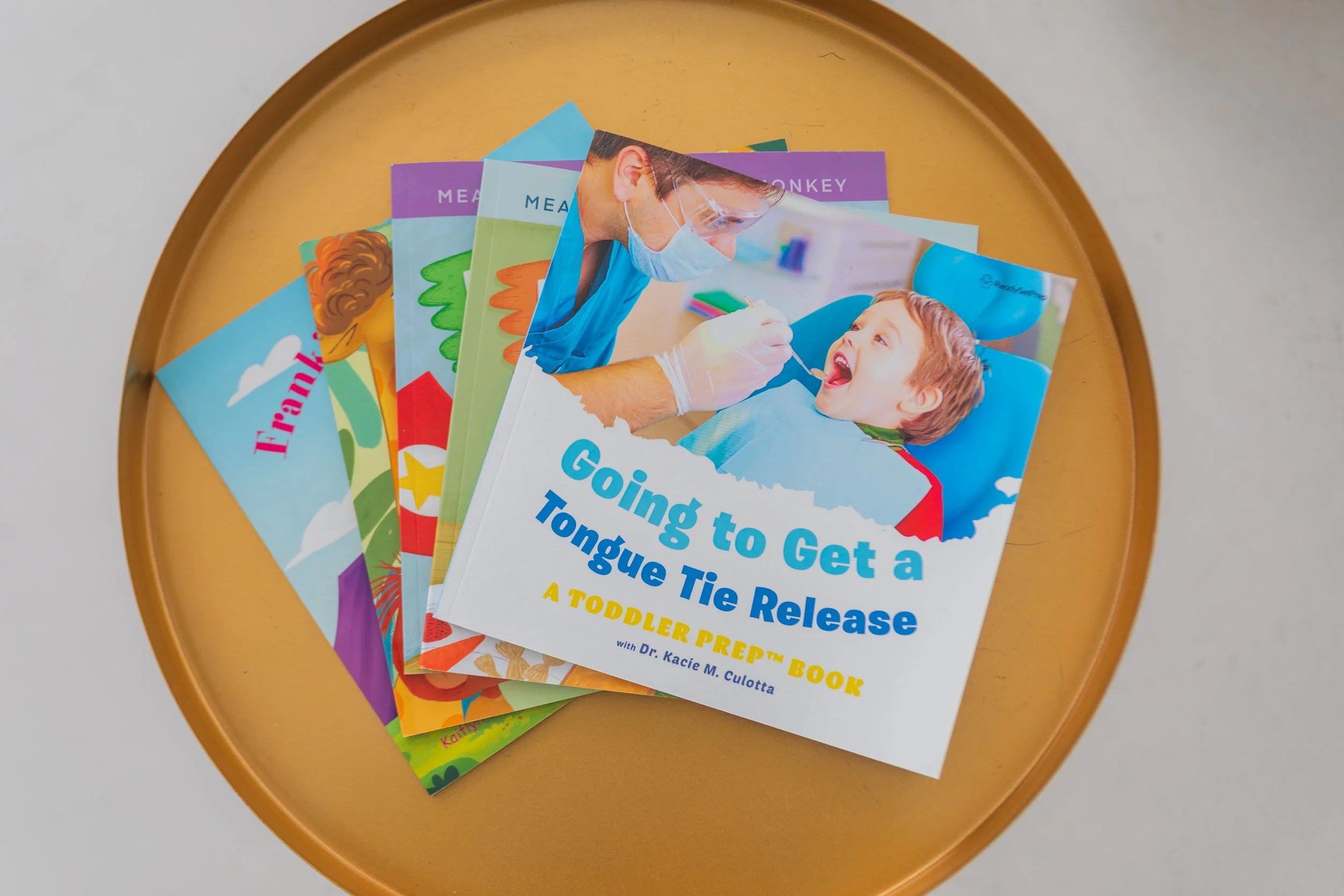 A stack of children's books, with the top book titled 'Going to Get a Tongue Tie Release: A Toddler Prep Book', featuring a child at the dentist with a dentist wearing a mask and gloves.