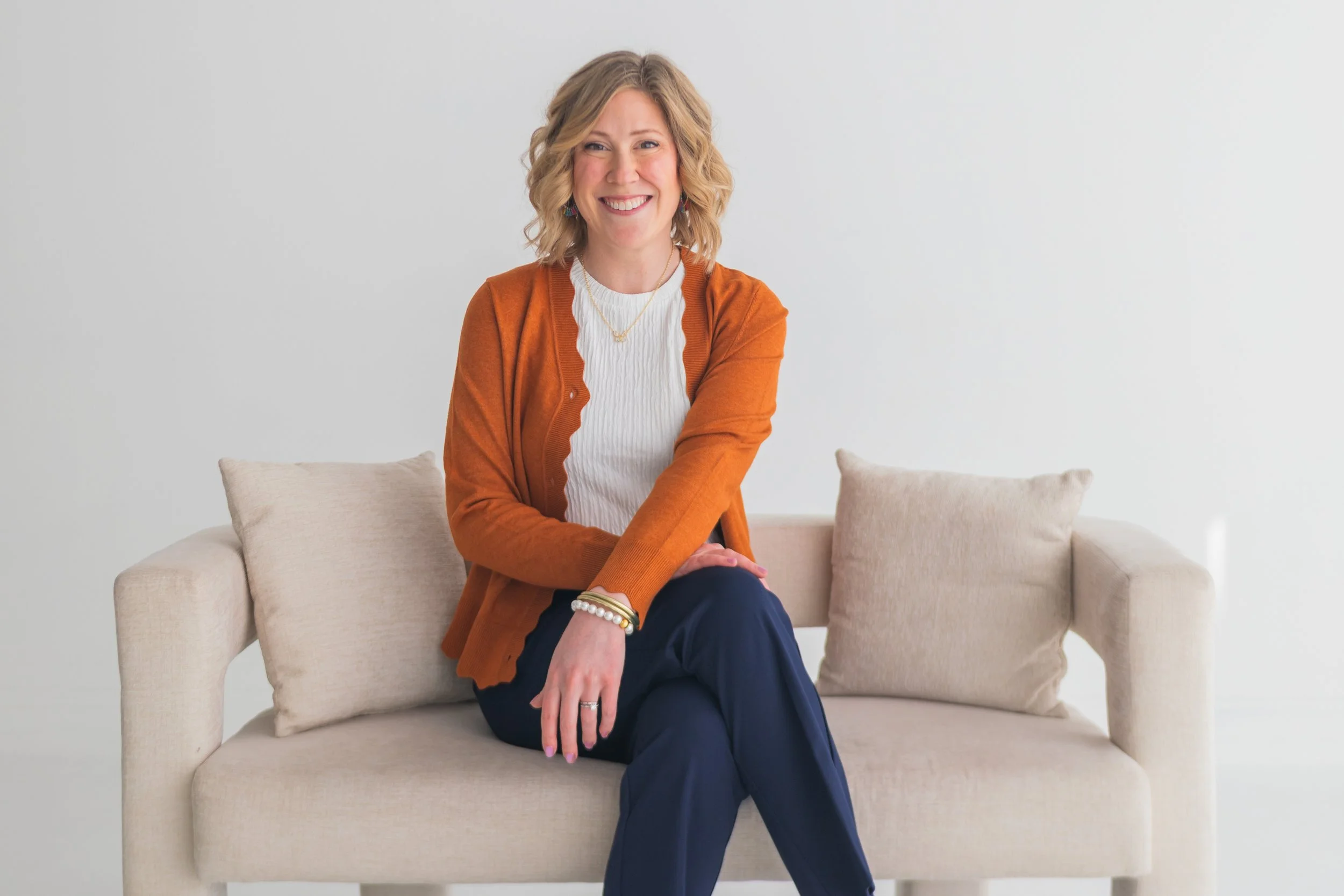 A woman with shoulder-length blonde curly hair smiling, sitting on a light-colored sofa with beige cushions in a minimalistic white room, wearing an orange cardigan, white top, and navy blue pants.