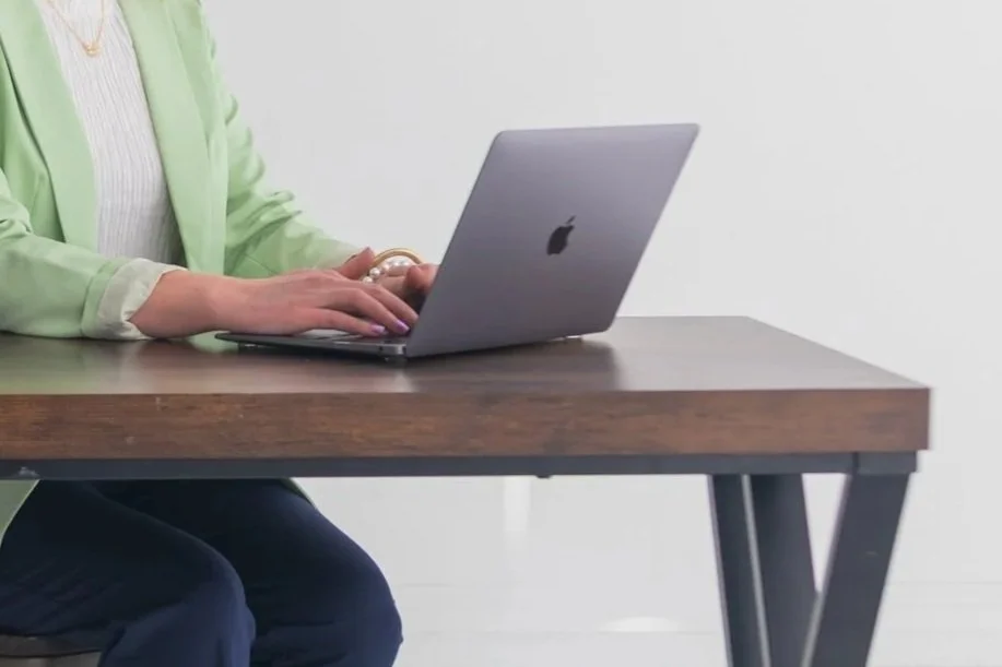 A person wearing a green blazer working on a silver Apple MacBook laptop at a wooden desk.