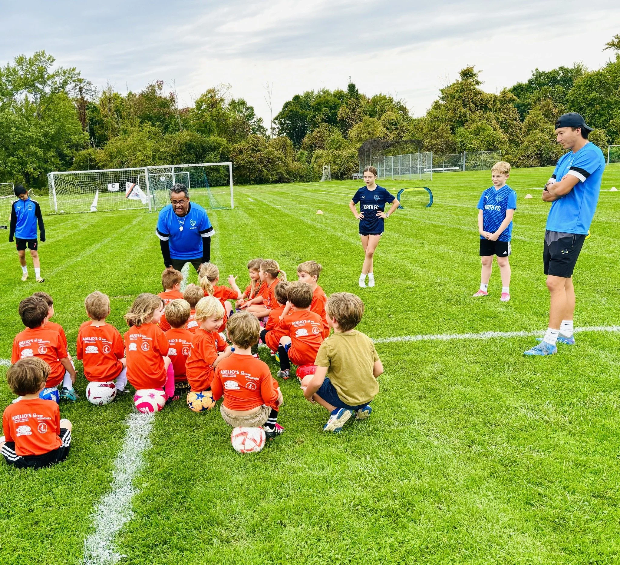 Children sitting on grass with soccer balls listening to coach on soccer field, coaches standing nearby, some kids in orange jerseys, others in blue, with goals and trees in background.