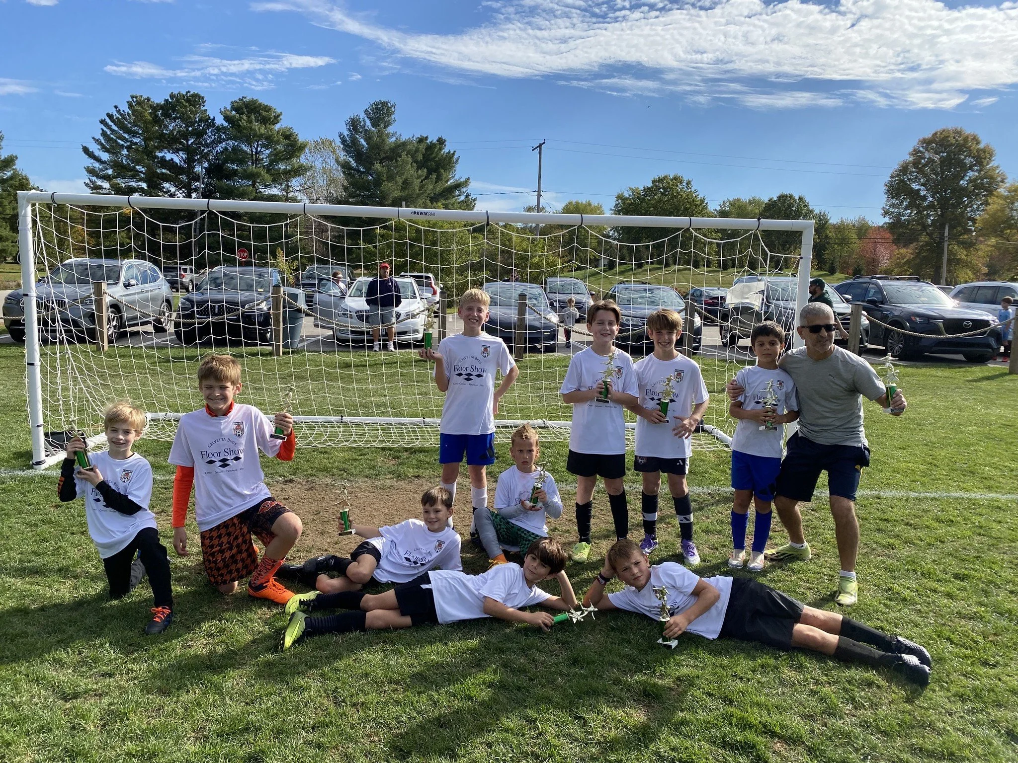 A youth soccer team with their coach posing after a game, holding trophies and drinks, on a grassy field with a soccer goal and parked cars in the background.