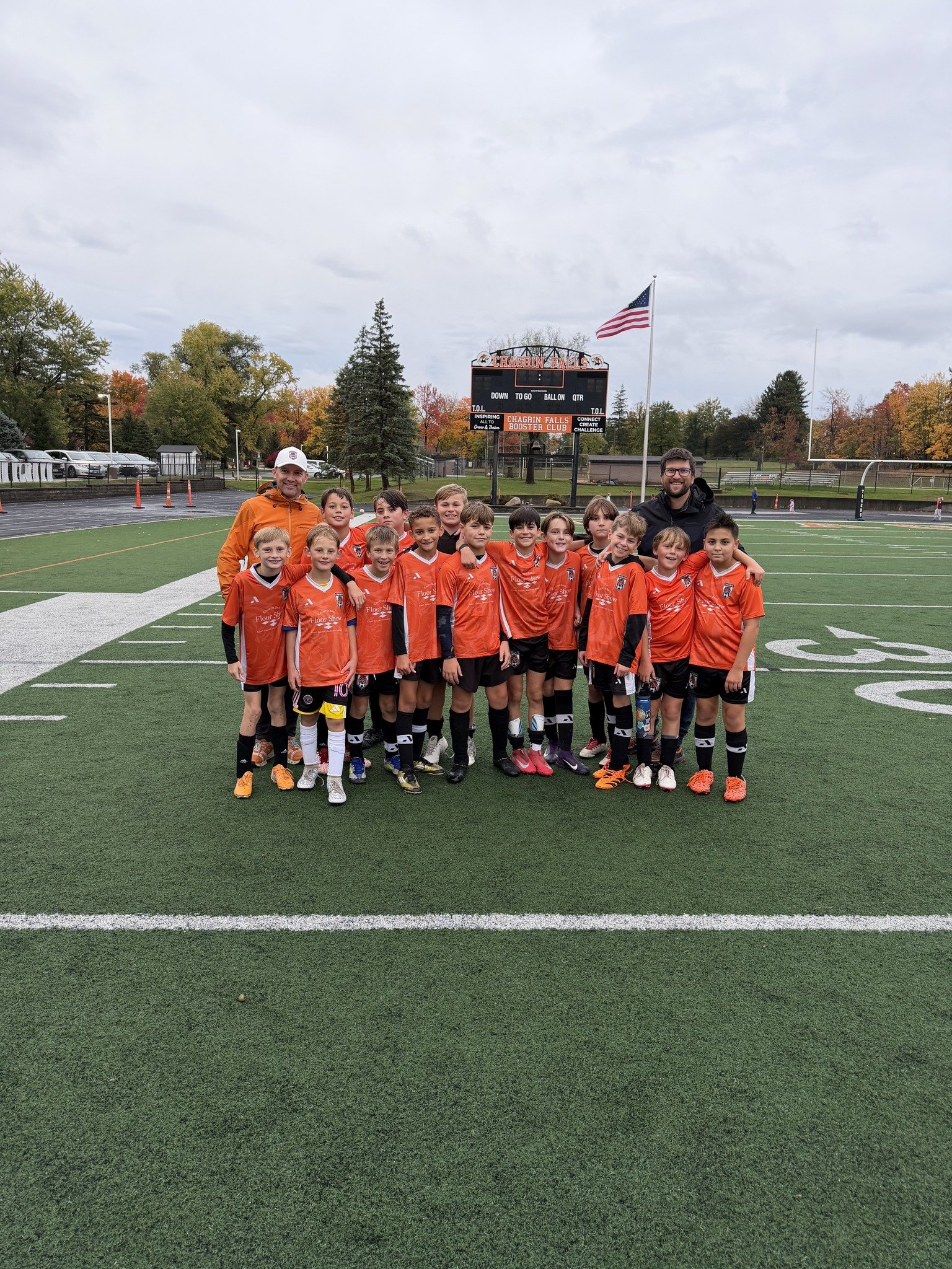 A youth soccer team in orange jerseys posing for a group photo on a football field, with two coaches and an American flag in the background.
