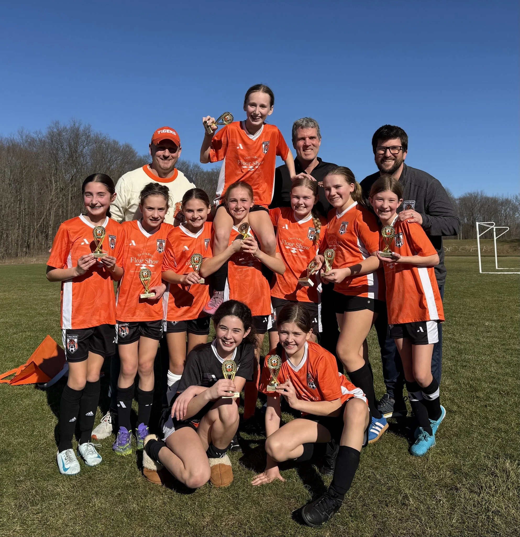 A youth girls' soccer team in orange jerseys celebrating with trophies on a sunny field, with coaches, one holding a small girl with a trophy on her shoulders.