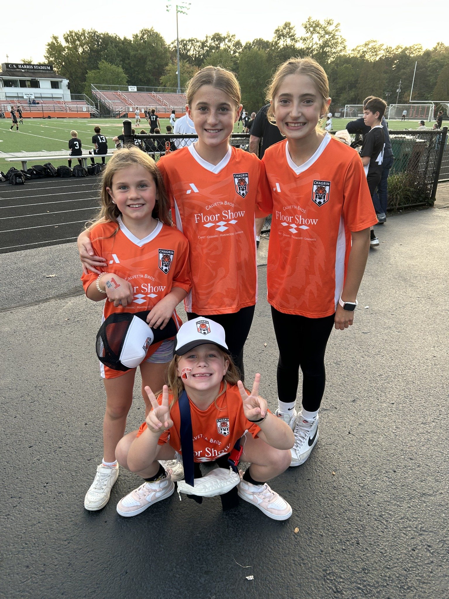 Four young girls in orange soccer jerseys posing for a picture on a soccer field, with a stadium and other players in the background.