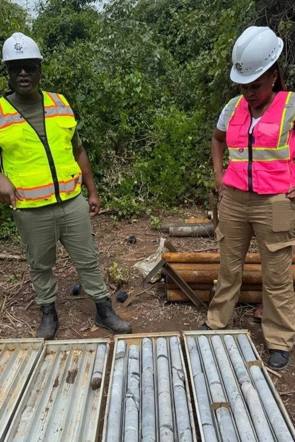 Two construction workers wearing safety helmets and high-visibility vests standing outdoors near metal pipes and equipment, with dense greenery in the background.