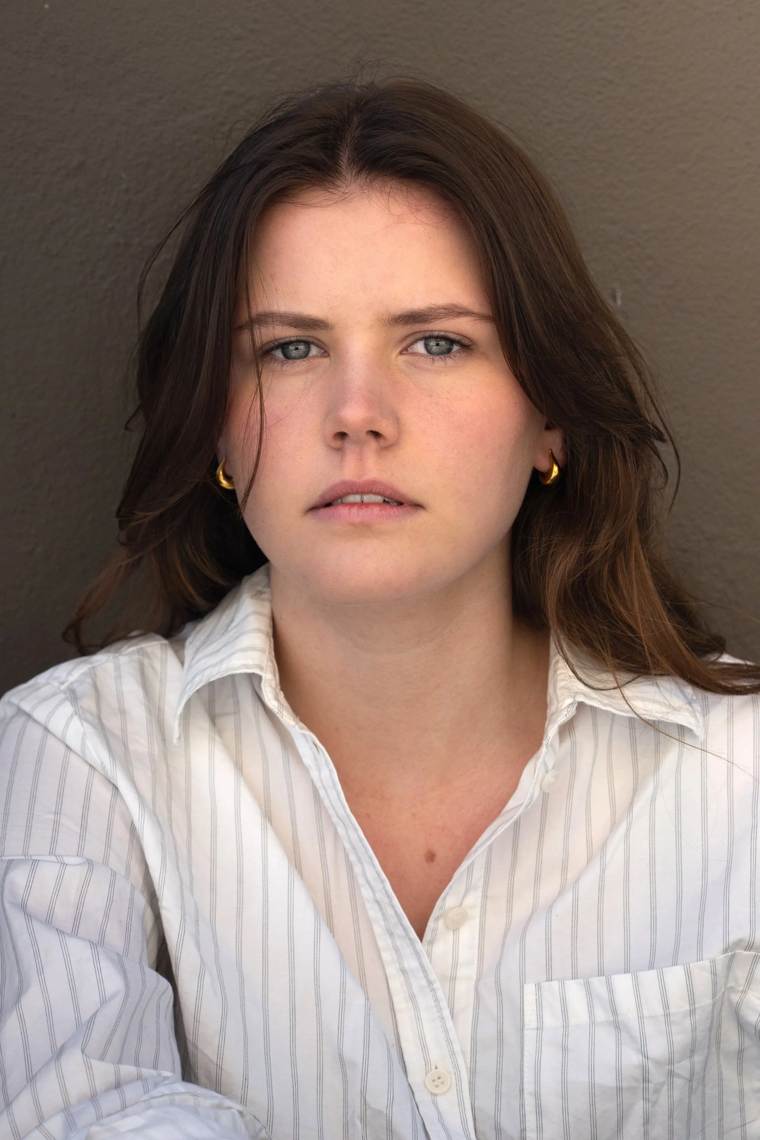 A young woman with shoulder-length brown hair, blue eyes, and wearing a white striped shirt with a collar. She is also wearing small gold hoop earrings. The background is a plain, textured wall.