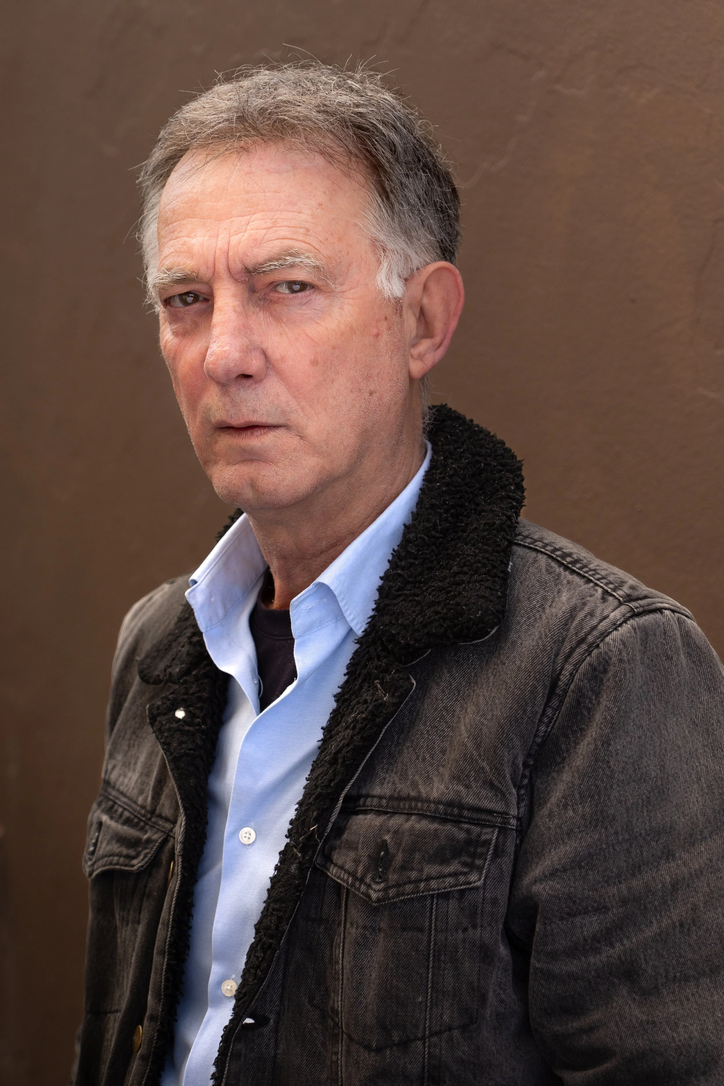 Close-up of a mature man with gray hair, wearing a denim jacket with a black shearling collar and a light blue shirt, standing against a plain brown background.