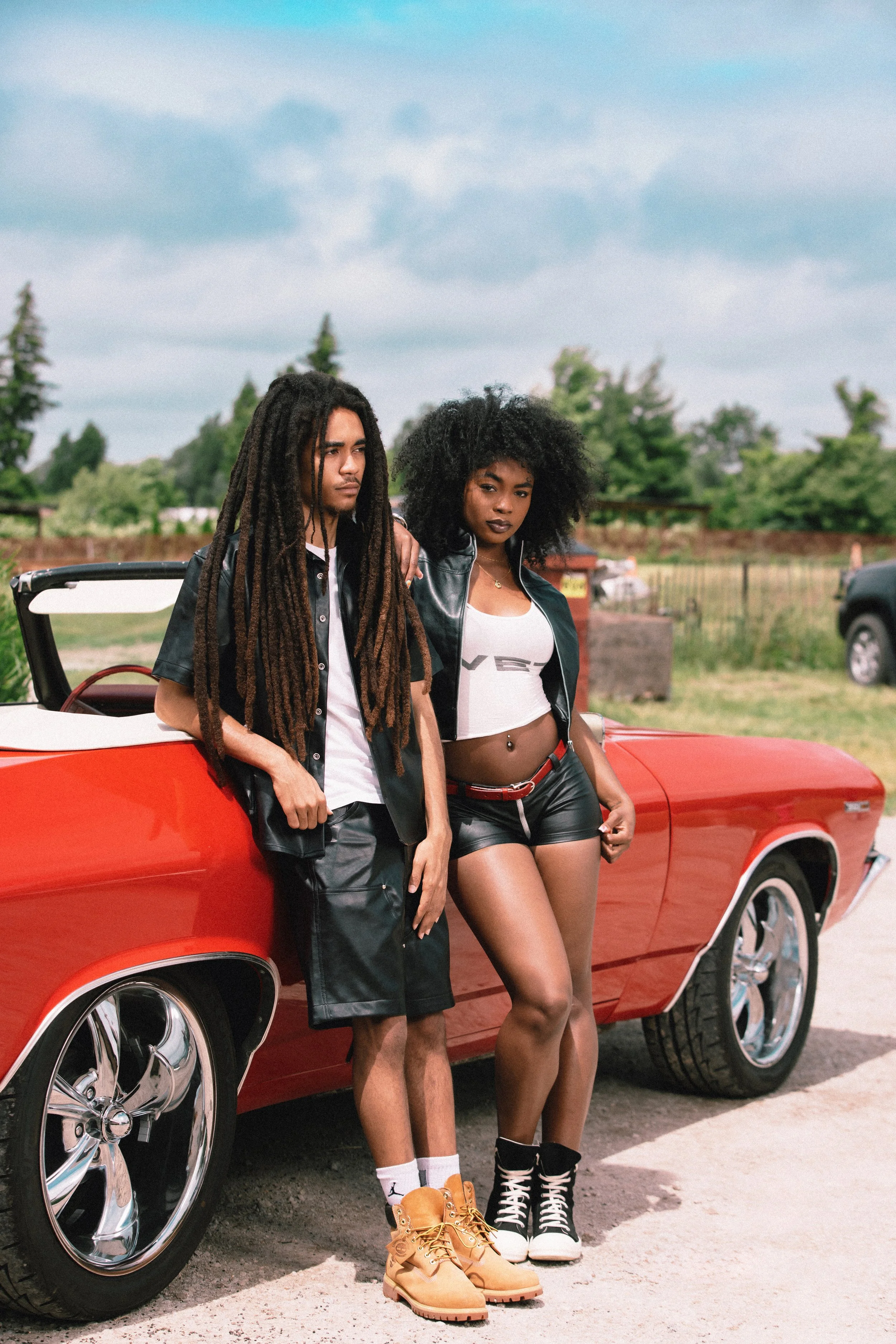 Two young people with dreadlocks and curly hair posing next to a red classic convertible car outdoors. The man is wearing a black shirt, shorts, white socks, and tan boots. The woman is dressed in a white crop top, black shorts, and high-top sneakers