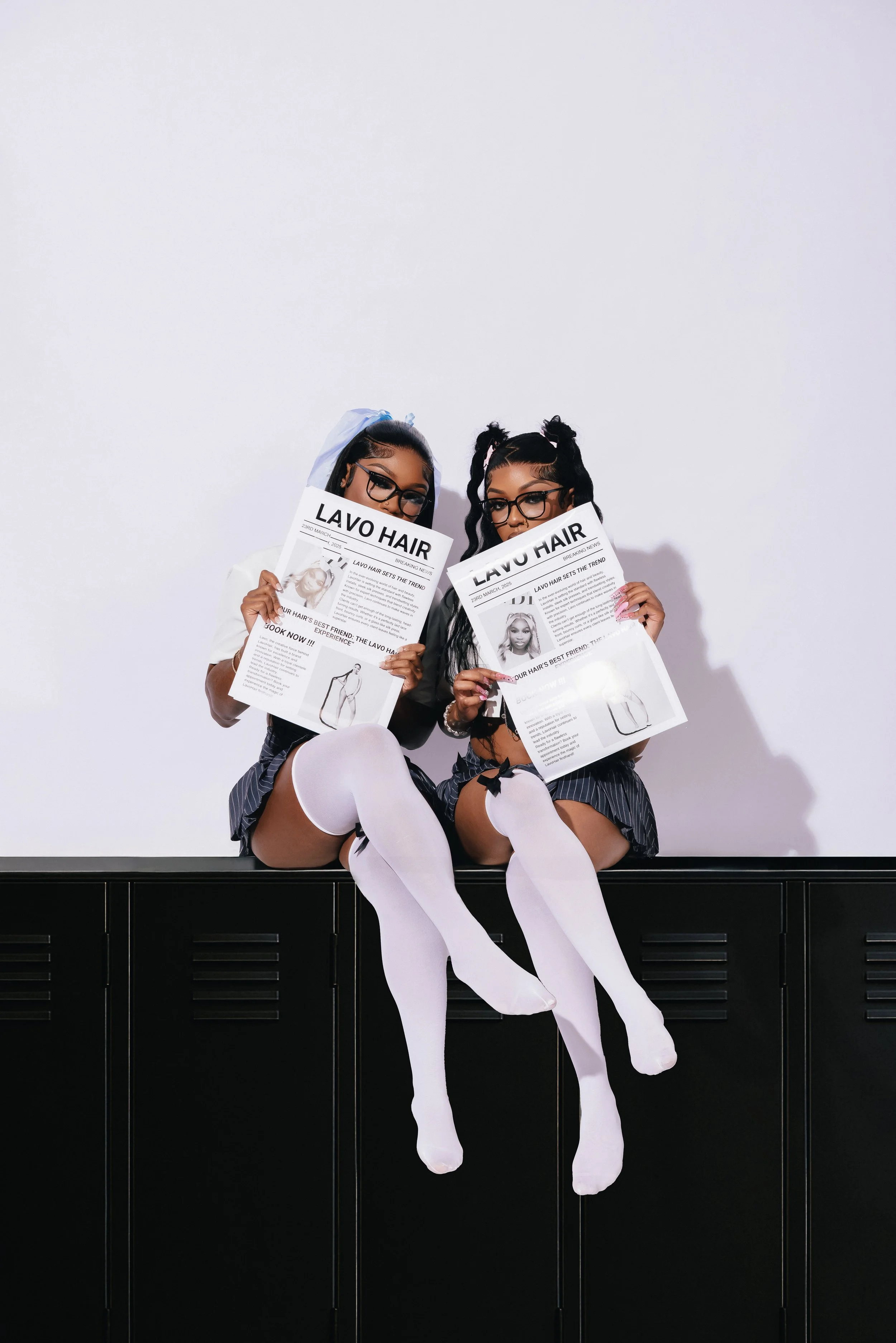 Two women dressed in schoolgirl costumes with white thigh-high socks and glasses, sitting on black lockers against a white wall, holding newspapers titled 'Lavo Hair' covering their faces.