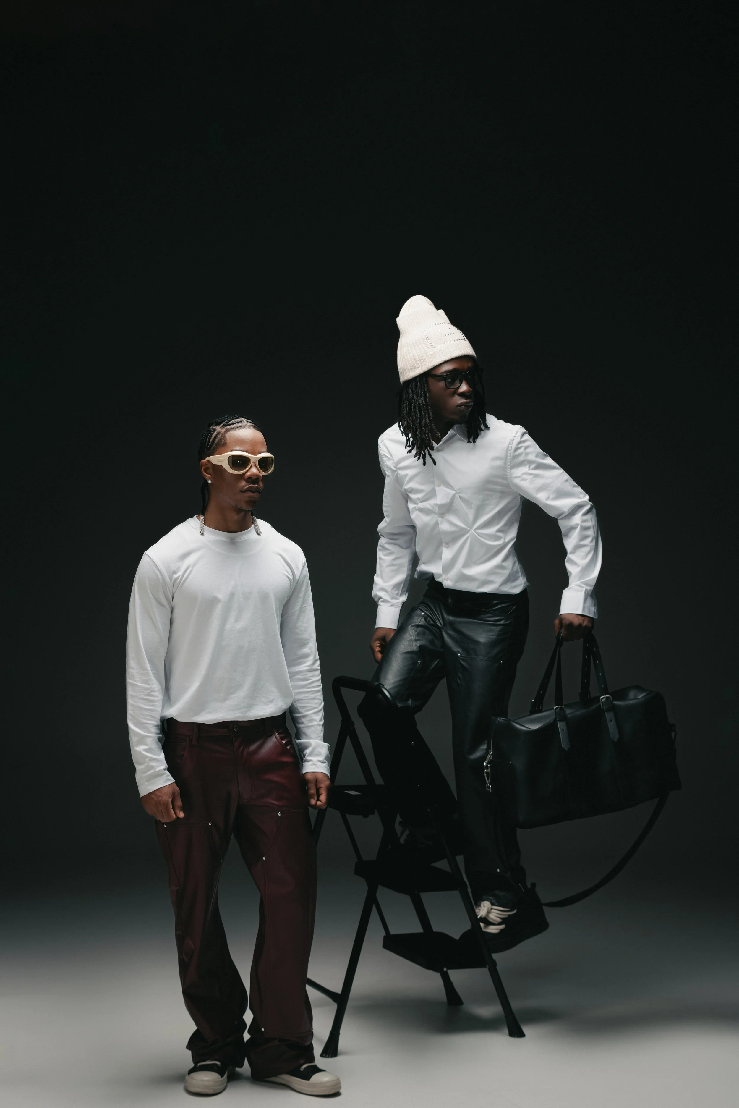 Two fashionably dressed young men, one standing with hands in pockets wearing sunglasses, the other on a step ladder holding a large bag, both in dark studio with black background.