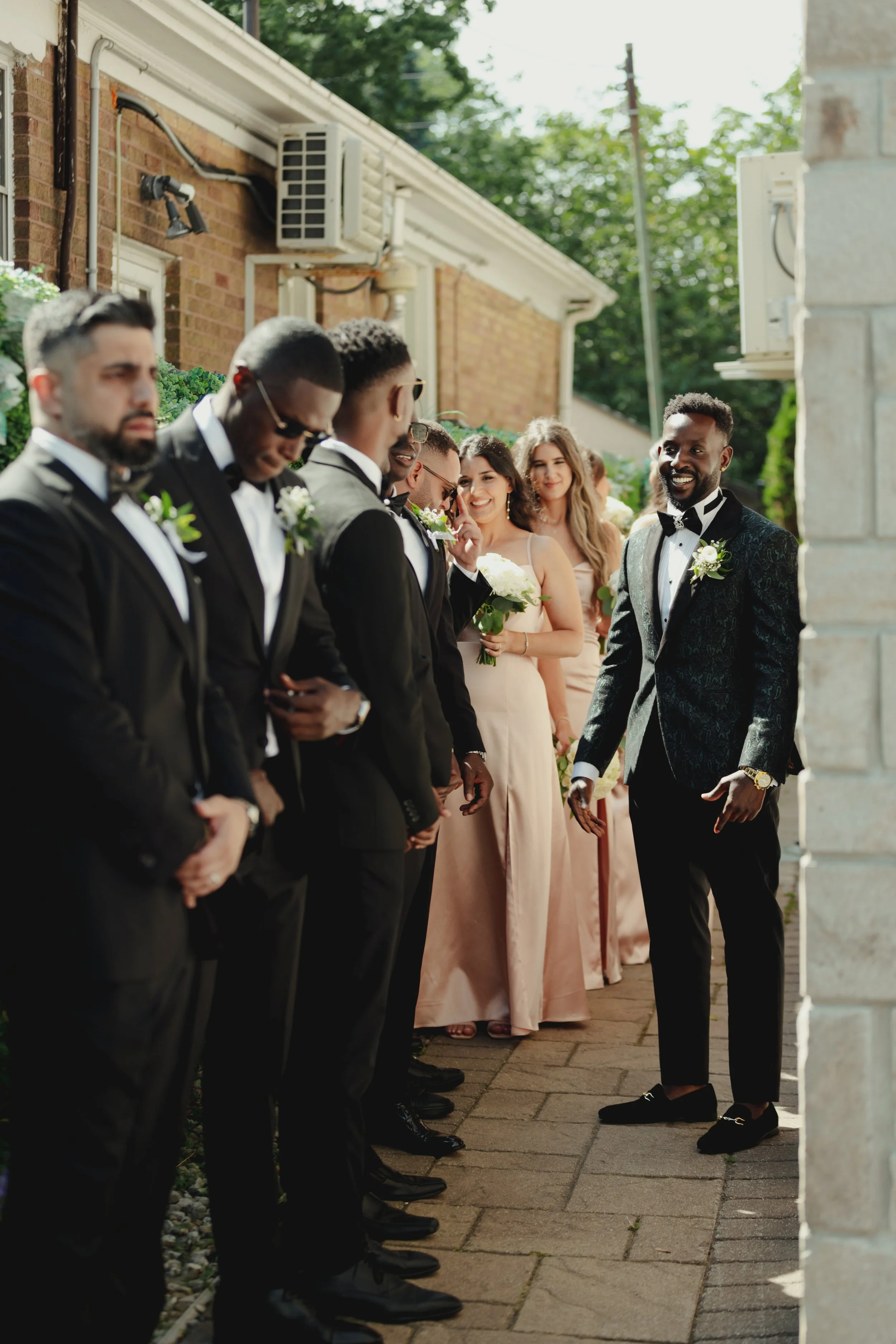 A groom and wedding party guests at an outdoor wedding ceremony, with the groom smiling, surrounded by friends and bridesmaids dressed in blush pink, during daytime.