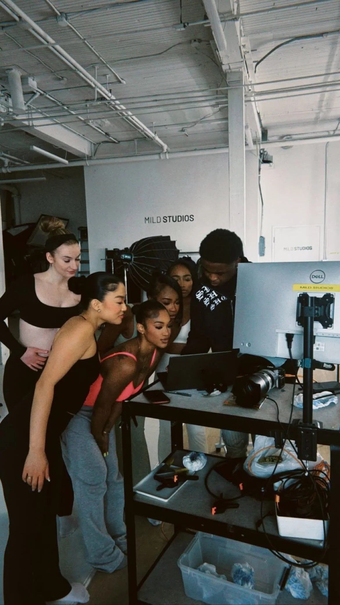 A group of six people, including five women and one man, gathered around a monitor in a photography or film studio, with equipment and lighting visible; they appear to be reviewing photos or footage together.