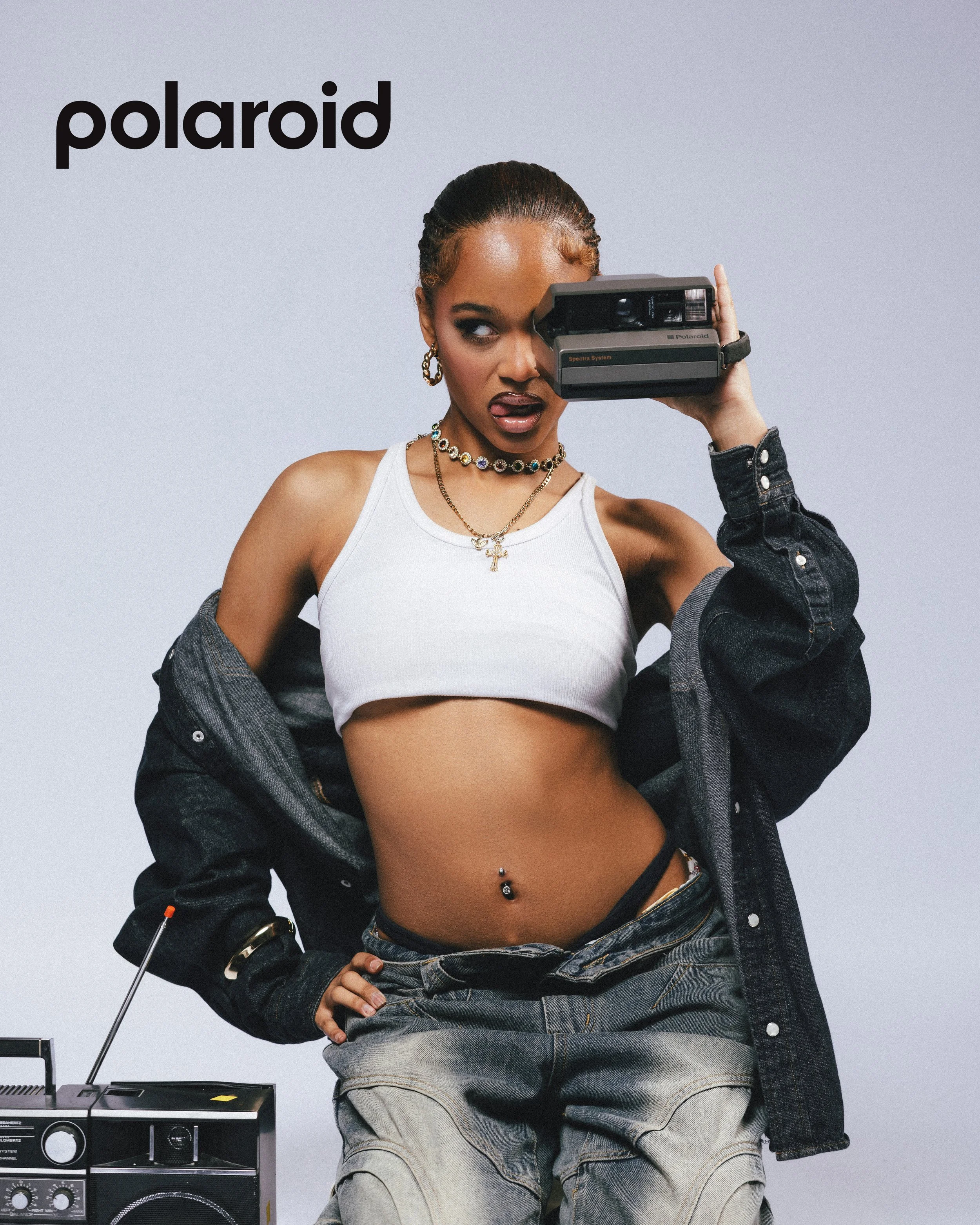 A young woman with a confident pose, holding a vintage Polaroid instant camera to her eye, sticking out her tongue with a playful expression. She has a belly button piercing, gold jewelry, and is wearing a white crop top, which is partly covered by a