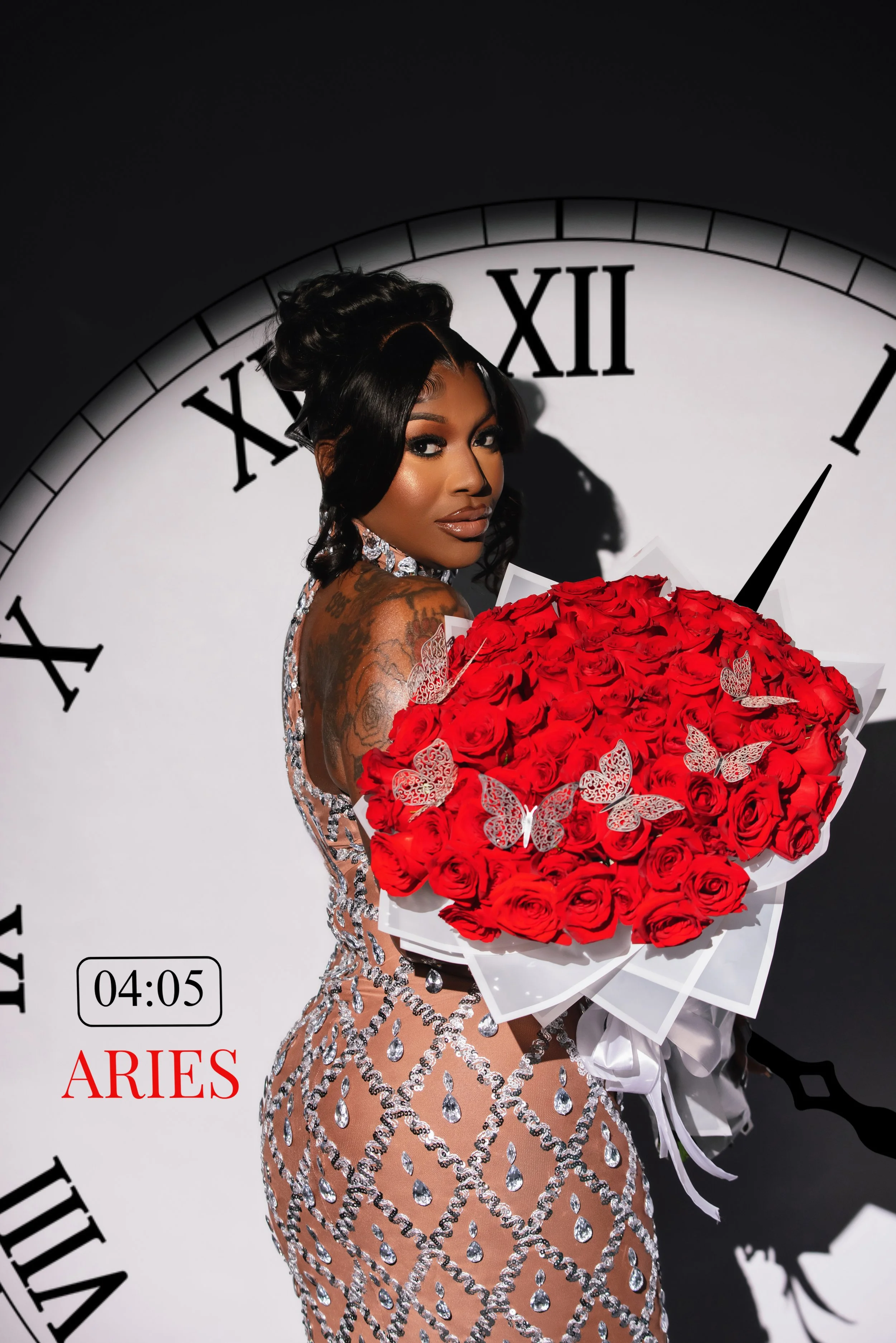 A woman with dark hair in an elegant updo, holding a large bouquet of red roses with decorative butterflies, standing in front of a large clock face showing 4:05, representing the Aries zodiac sign.