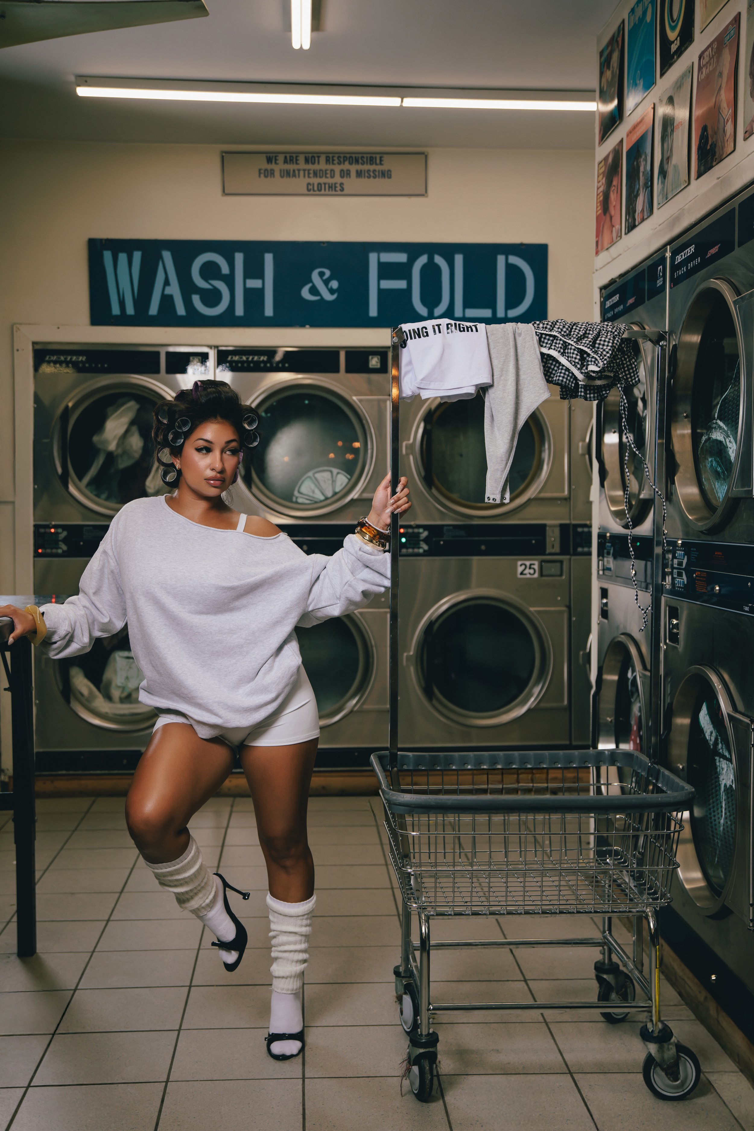 A young woman with hair rollers in her hair stands in a laundromat holding a sign that reads 'Doing it right.' She is wearing a loose gray sweatshirt, white shorts, and socks with sandals. There are washing machines behind her and laundry hanging on 