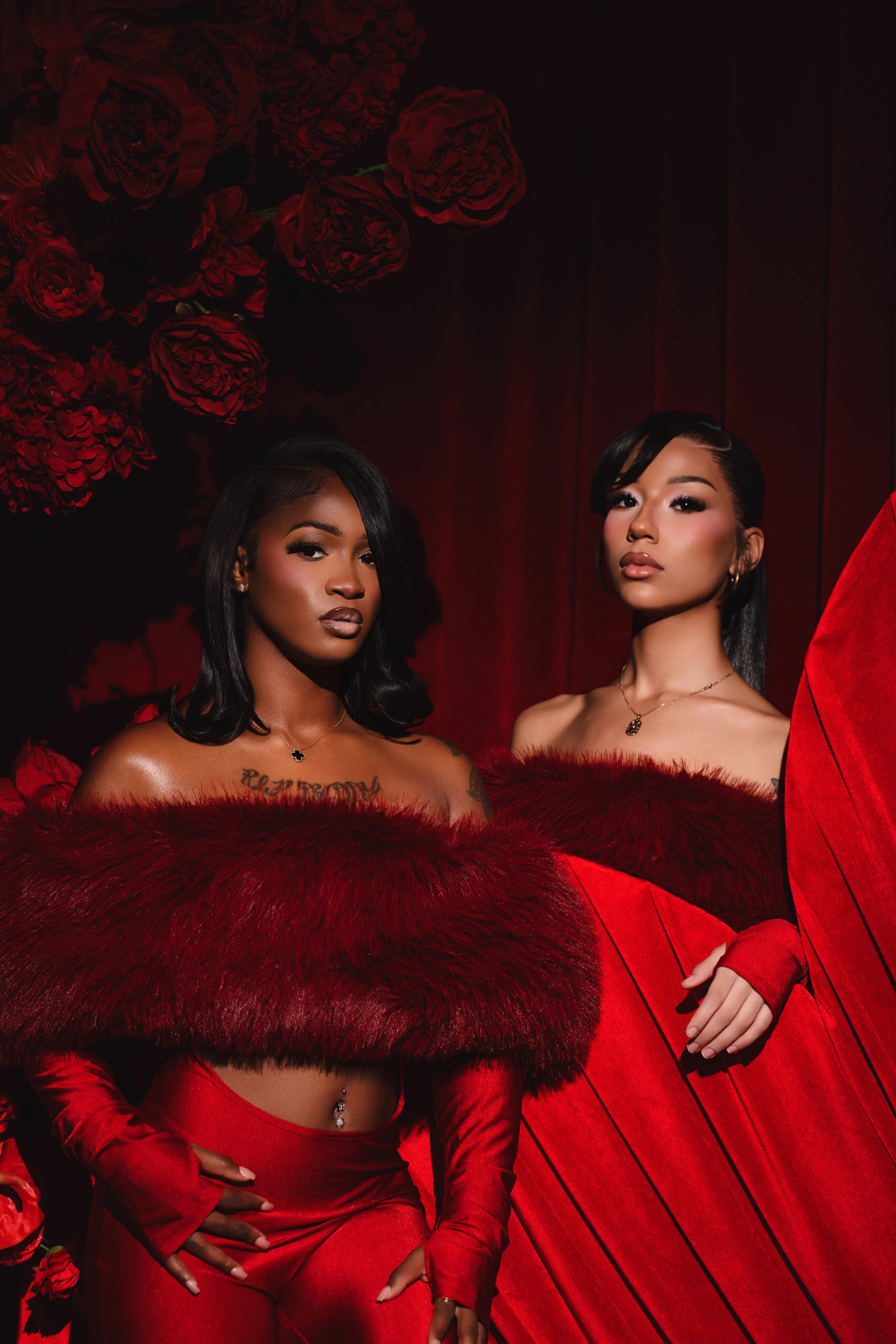 Two women dressed in red, standing against a backdrop of red flowers and curtains, with confident expressions.
